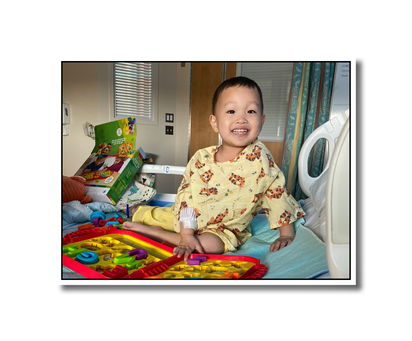 A younger Noah smiling at the camera. He's sitting on his hospital bed, wearing tigers on his gown and playing with toys.