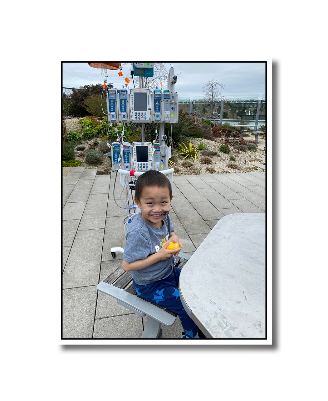 Noah smiling and enjoying a snack in the courtyard of the hospital. He has an array of IV pumps and machines in tow.