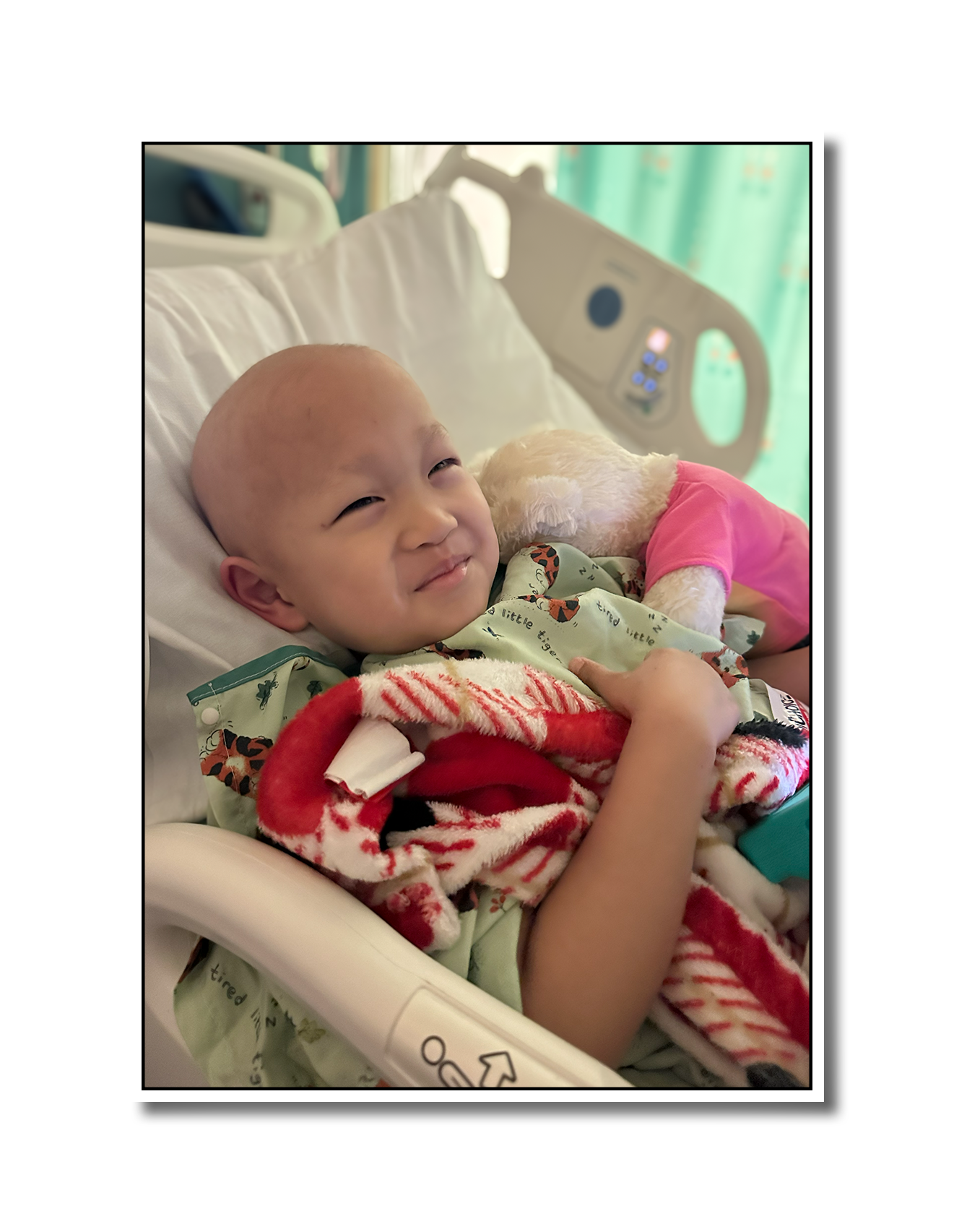 A young child in a hospital bed smiling and lying with a stuffed animal, surrounded by hospital equipment and green curtains in the background.