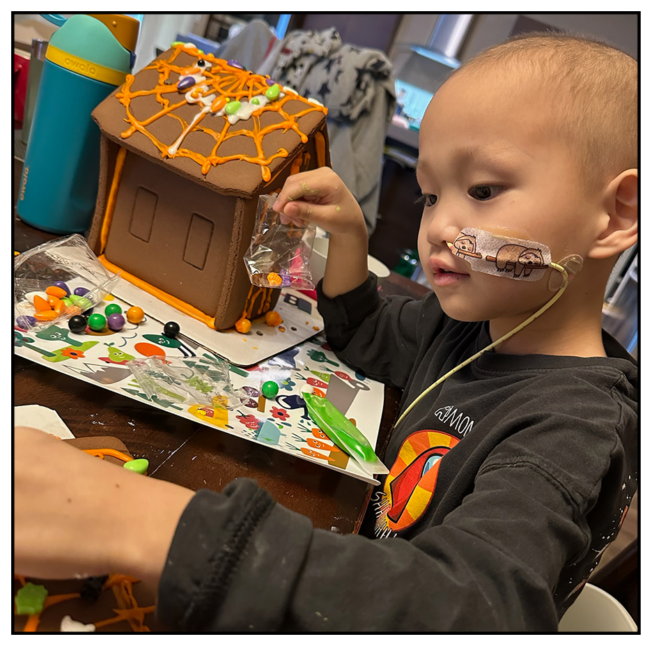 A young boy with a bandage on his face is decorating a gingerbread house with colorful candies. The gingerbread house has orange icing on the roof and is on a festive holiday-themed table.