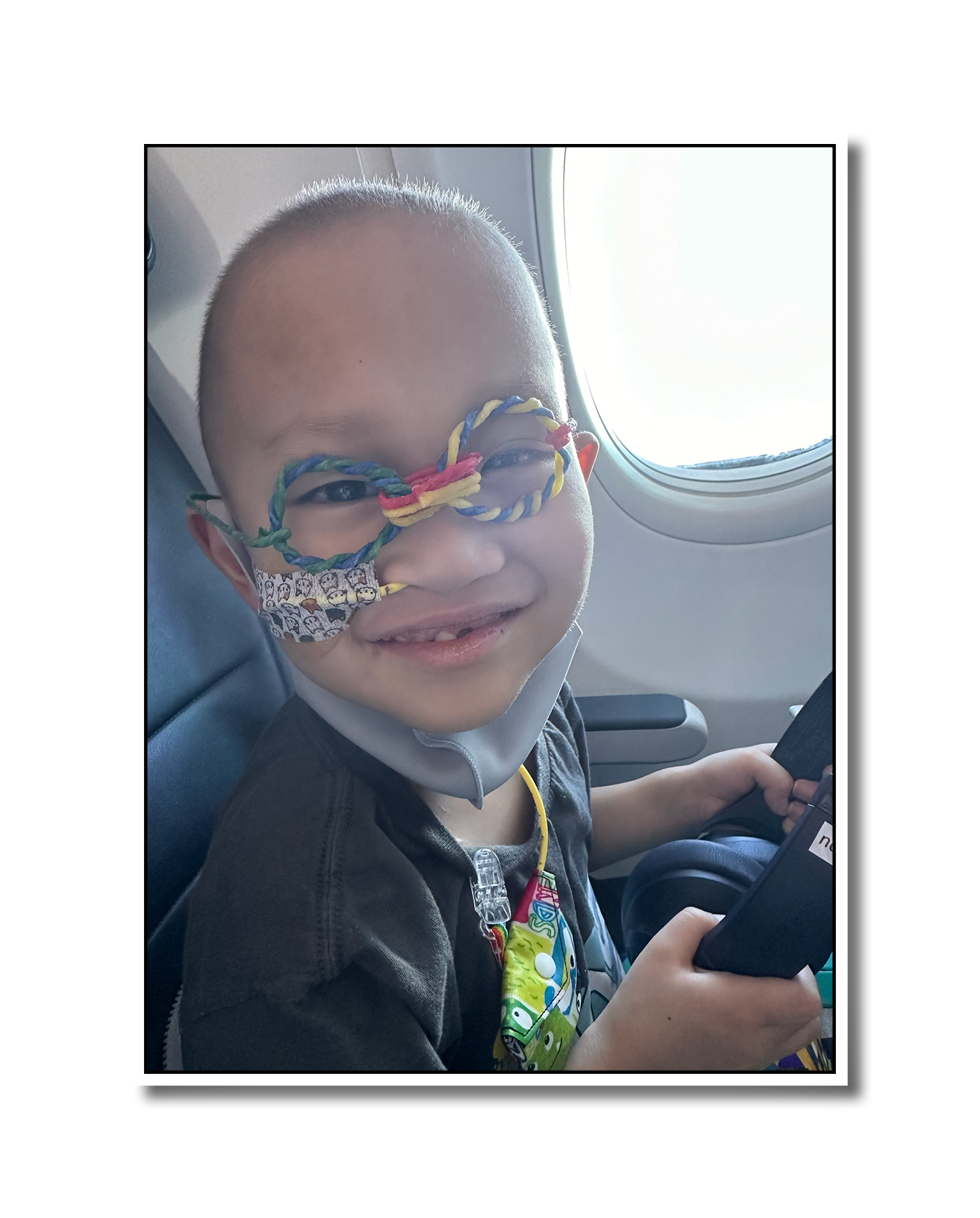 Noah smiling for the camera from his airplane seat. He's holding headphones in his hands, and has goofy fake glasses on his face. His hair is light and fuzzy from current chemo, and has a feeding tube in his nose.