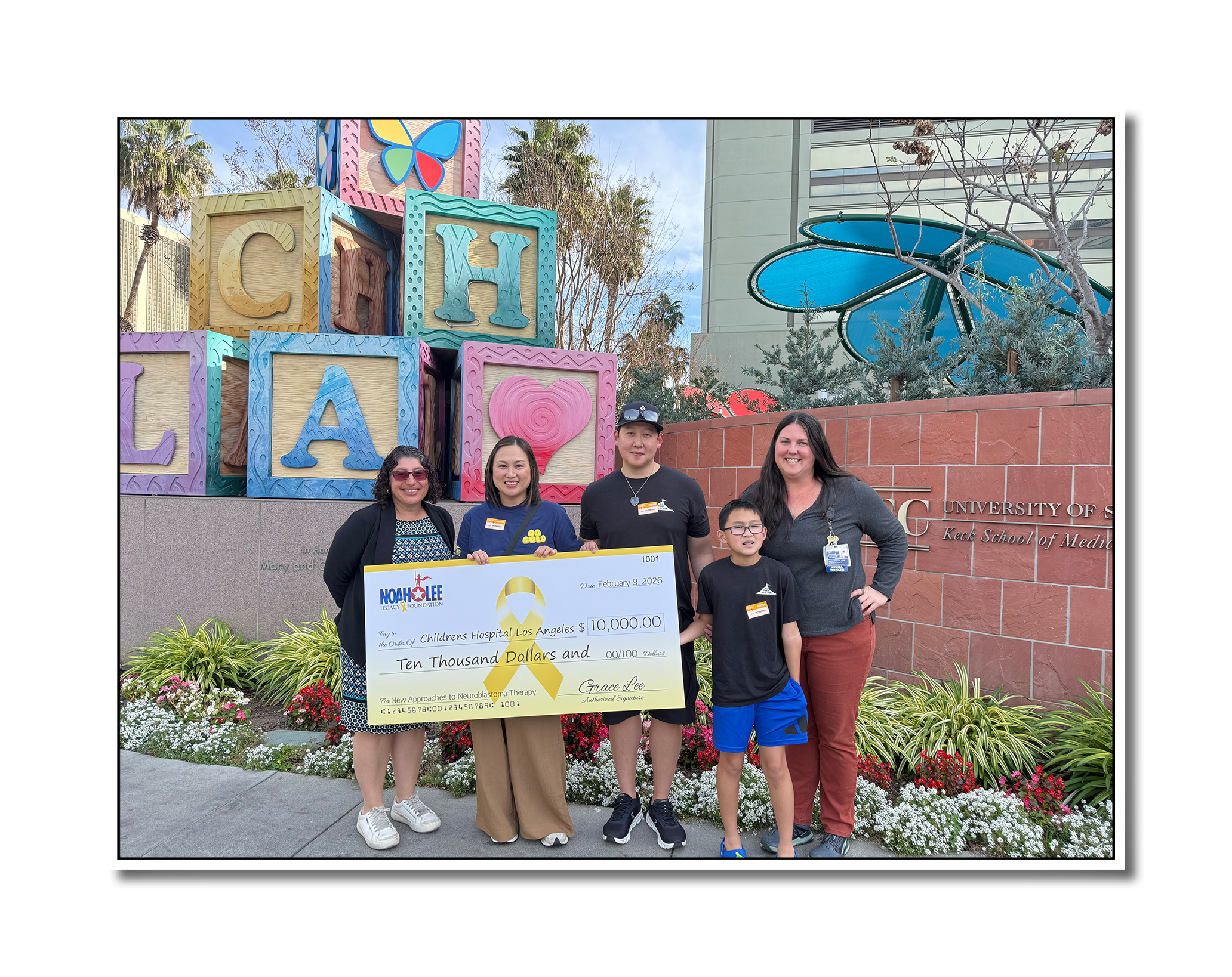 Group of people standing in front of a colorful Children's Hospital Los Angeles sign holding a large check for $10,000 for neuroblastoma therapy research at the University of Southern California, with flowers and a building in the background.