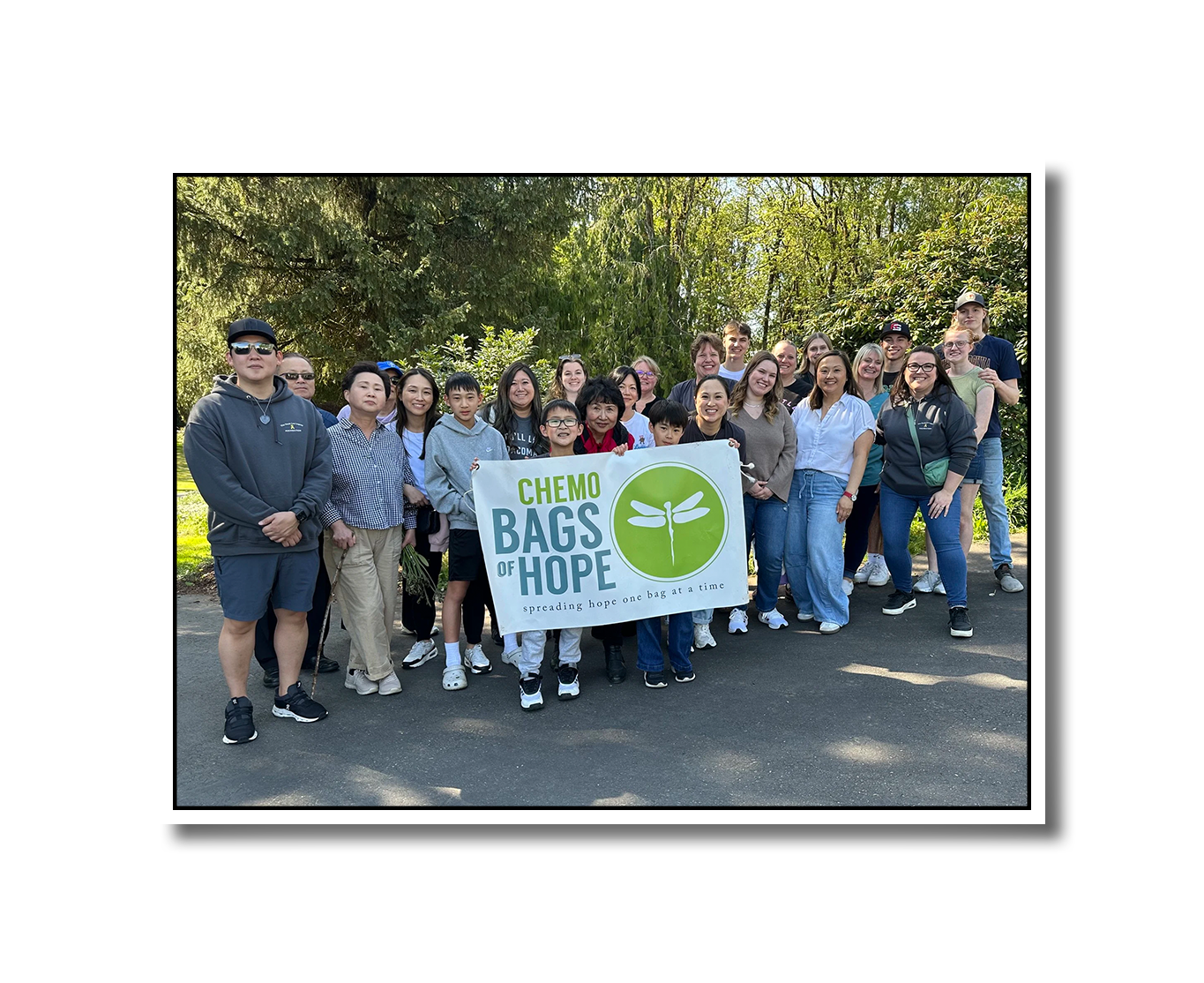 Group of people outdoors holding a banner that reads 'Chemo Bags of Hope' with a logo of a dragonfly and the tagline 'spreading hope one bag at a time', surrounded by trees and sunlight.
