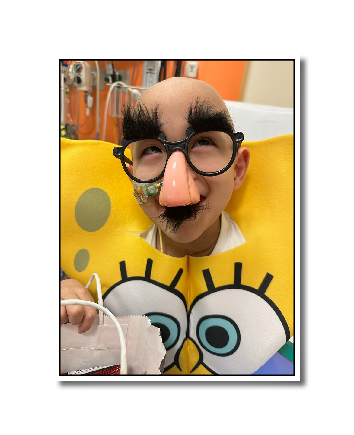 A person dressed as SpongeBob SquarePants with a large yellow sponge costume, big eyes, and a fake nose, wearing glasses and a mustache, in a hospital room.
