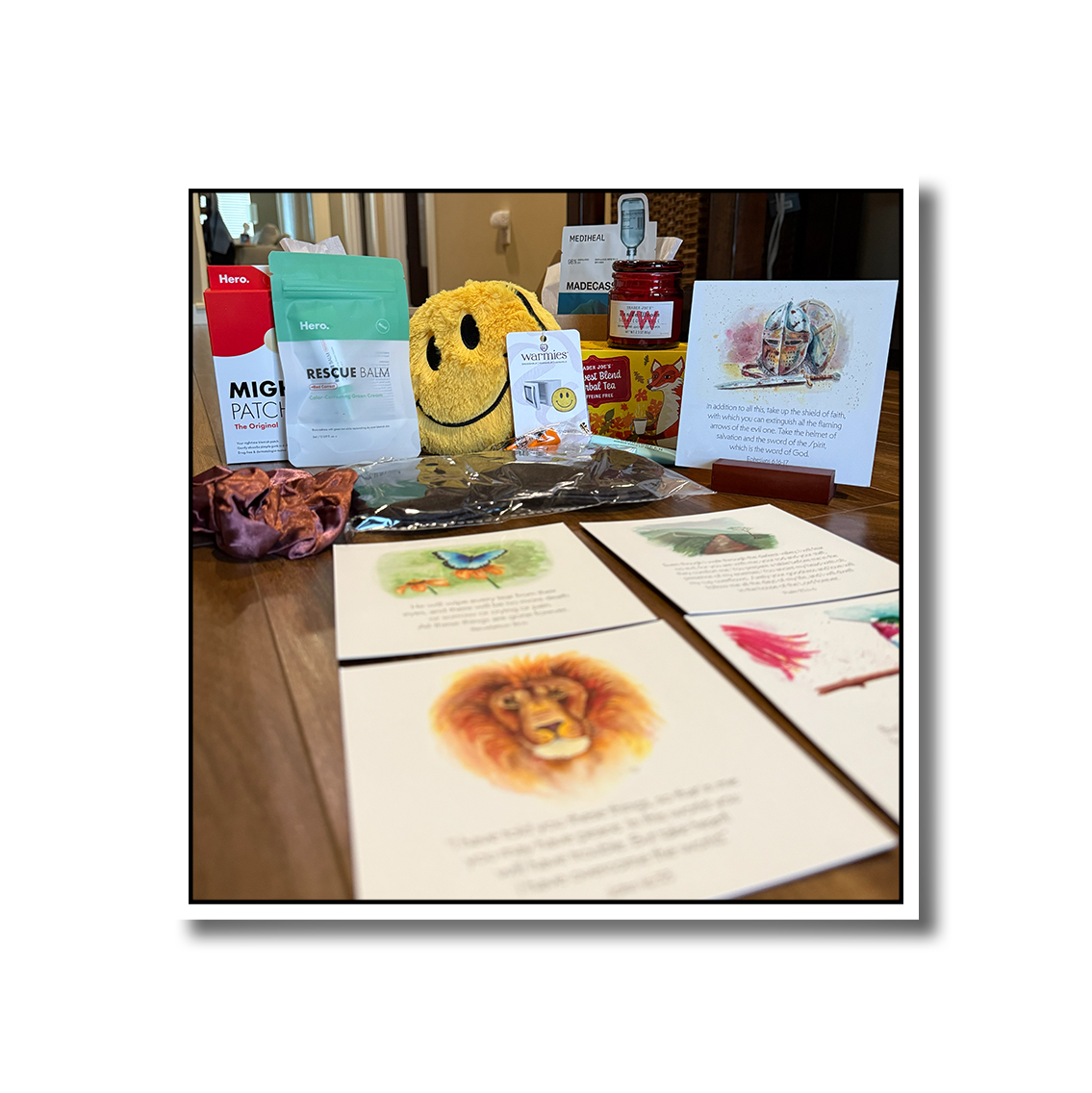 A photo of care package contents, including pimple patches, a smiley face warmie, candle, scrunchie, and hand painted artwork featuring Bible verses.