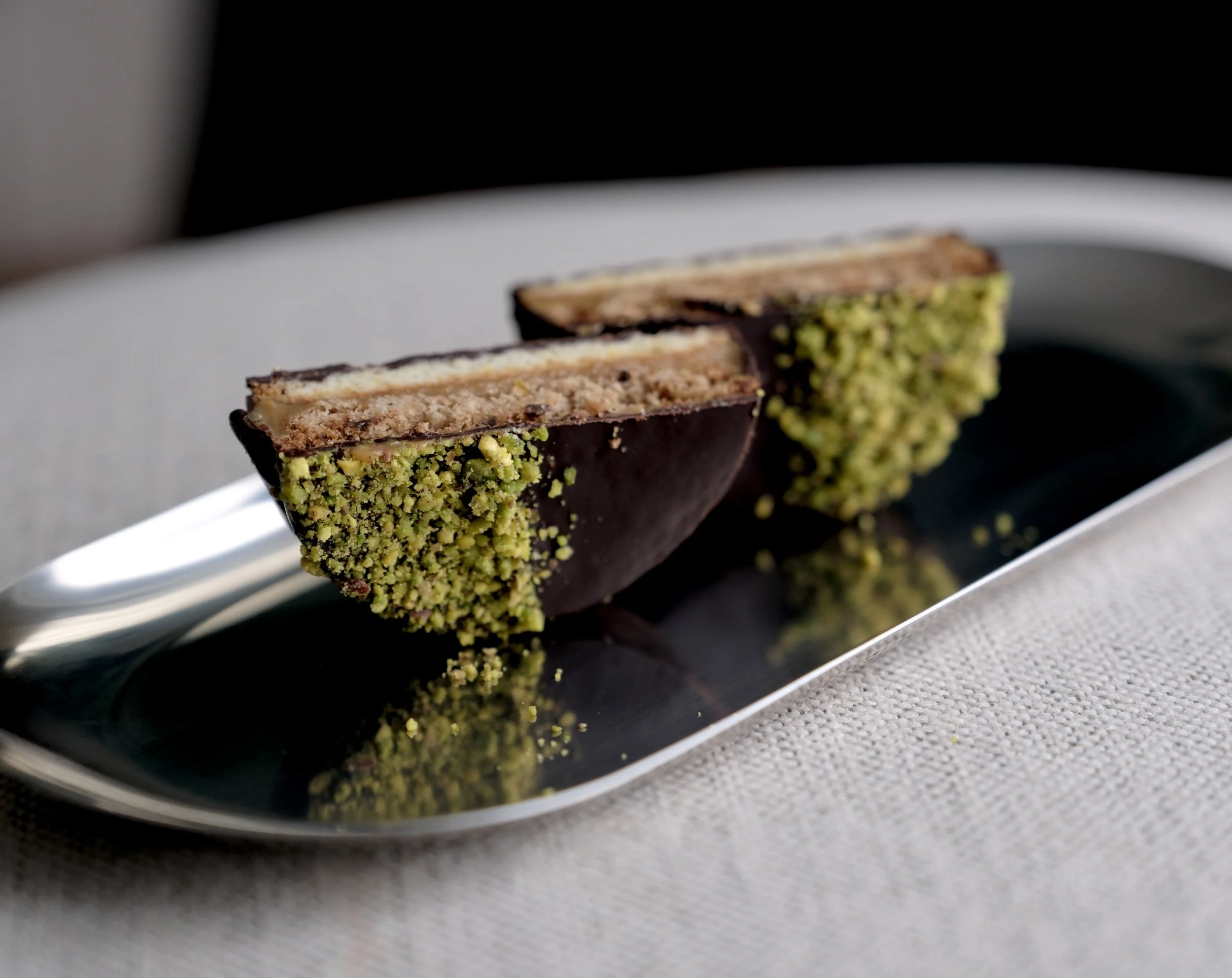 Two slices of layered dessert dipped in dark chocolate and coated with crushed pistachios, served on a silver tray.