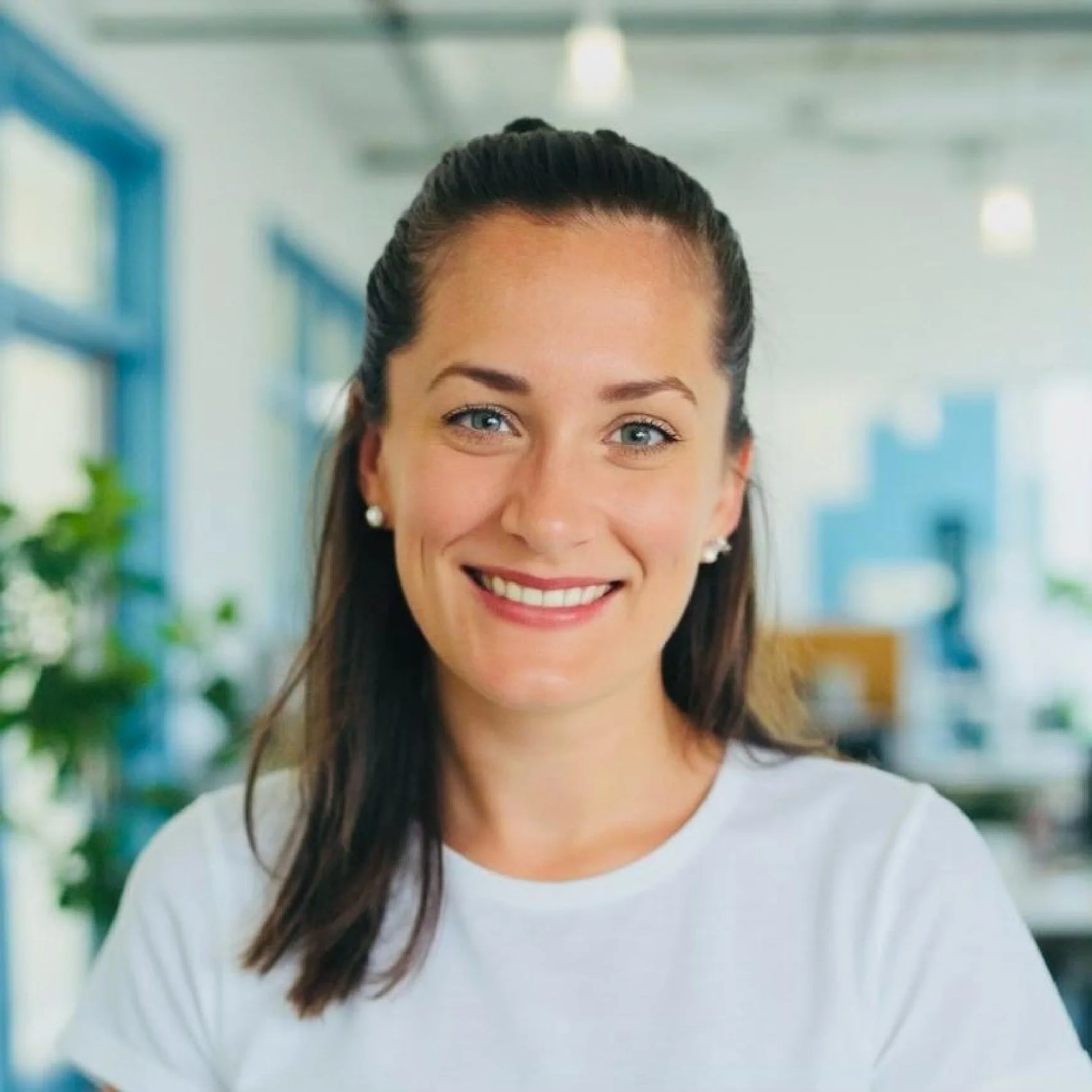 Smiling woman with long dark hair, blue eyes, and pearl earrings in a bright indoor space with blurred background of blue and white.