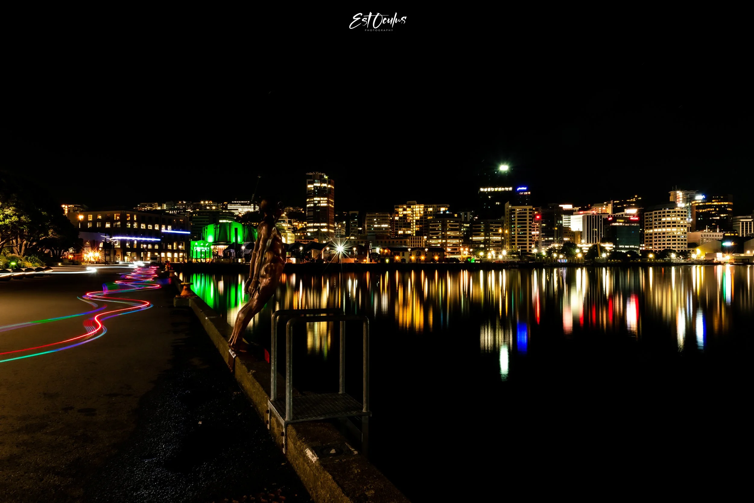 Night view of a city skyline with colorful light reflections in water and light trails on a street, featuring a statue in the foreground.
