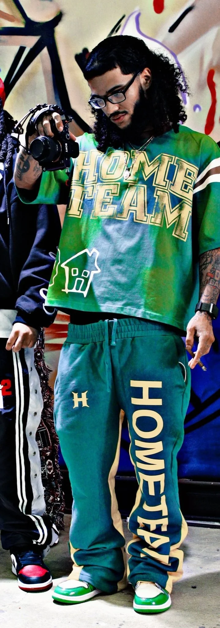 Young man with dark curly hair, glasses, and tattoos, wearing a green and blue 'HOMIE TEAM' t-shirt and green jogger pants with 'HOMIE TEAM' printed on them, holding a camera with a strap, standing in front of a colorful graffiti wall.