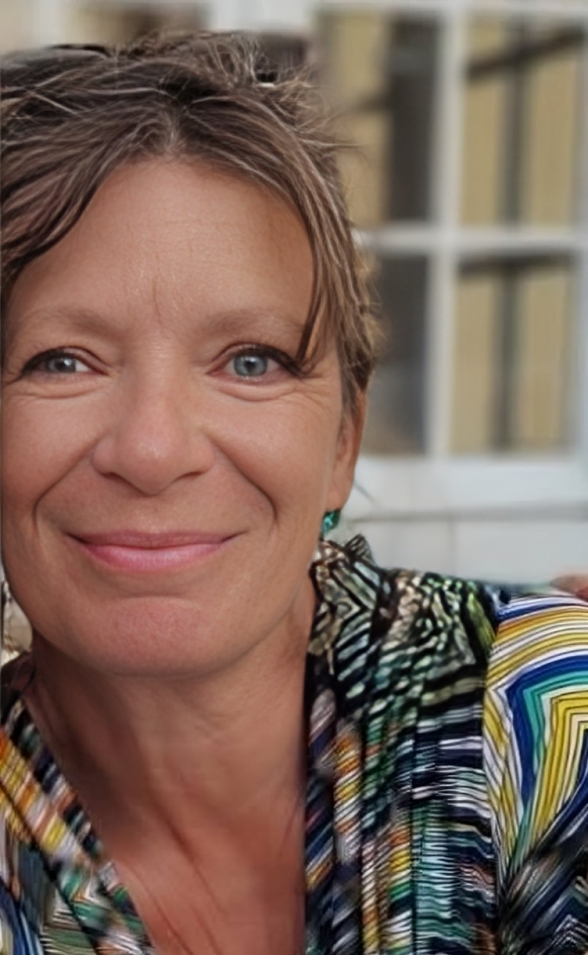 Une femme souriante avec des cheveux bruns et un haut coloré, en arrière-plan flou d'une fenêtre.