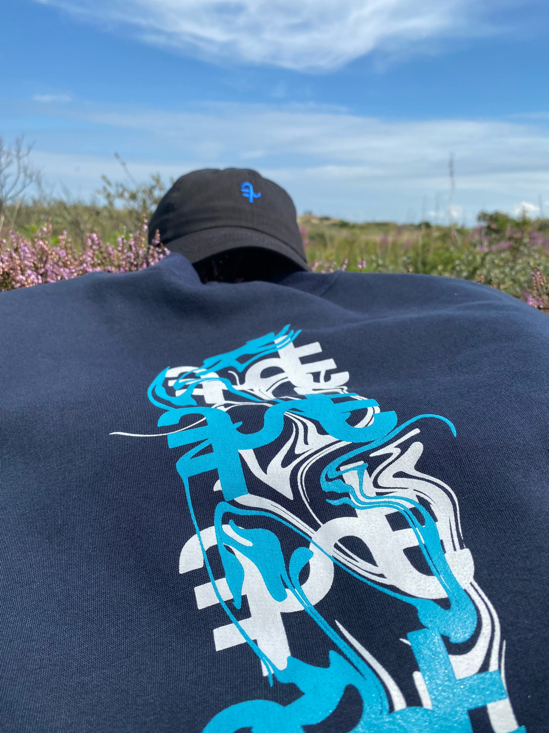 Person wearing a navy blue shirt with abstract blue and white graphics, lying on grass with purple flowers, under a blue sky.