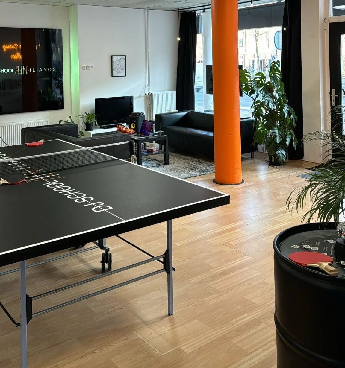 Interior of a modern room with a black ping pong table, black couch, orange pillar, plants, and a TV on a stand.