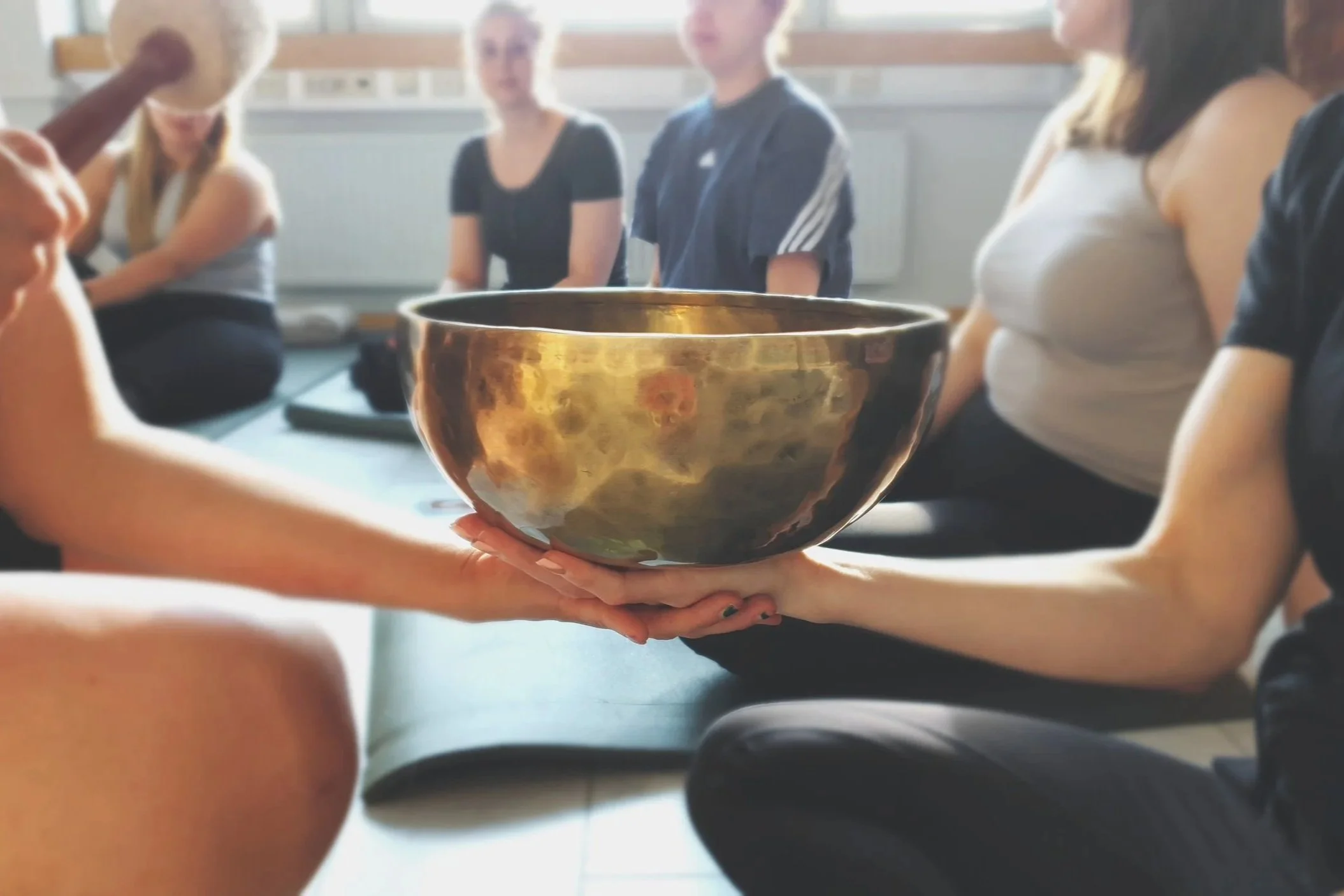 Kuvassa ihmisiä istumassa sisätiloissa, ja etummaisena kädessä pidetään kullanväristä äänimaljaa.