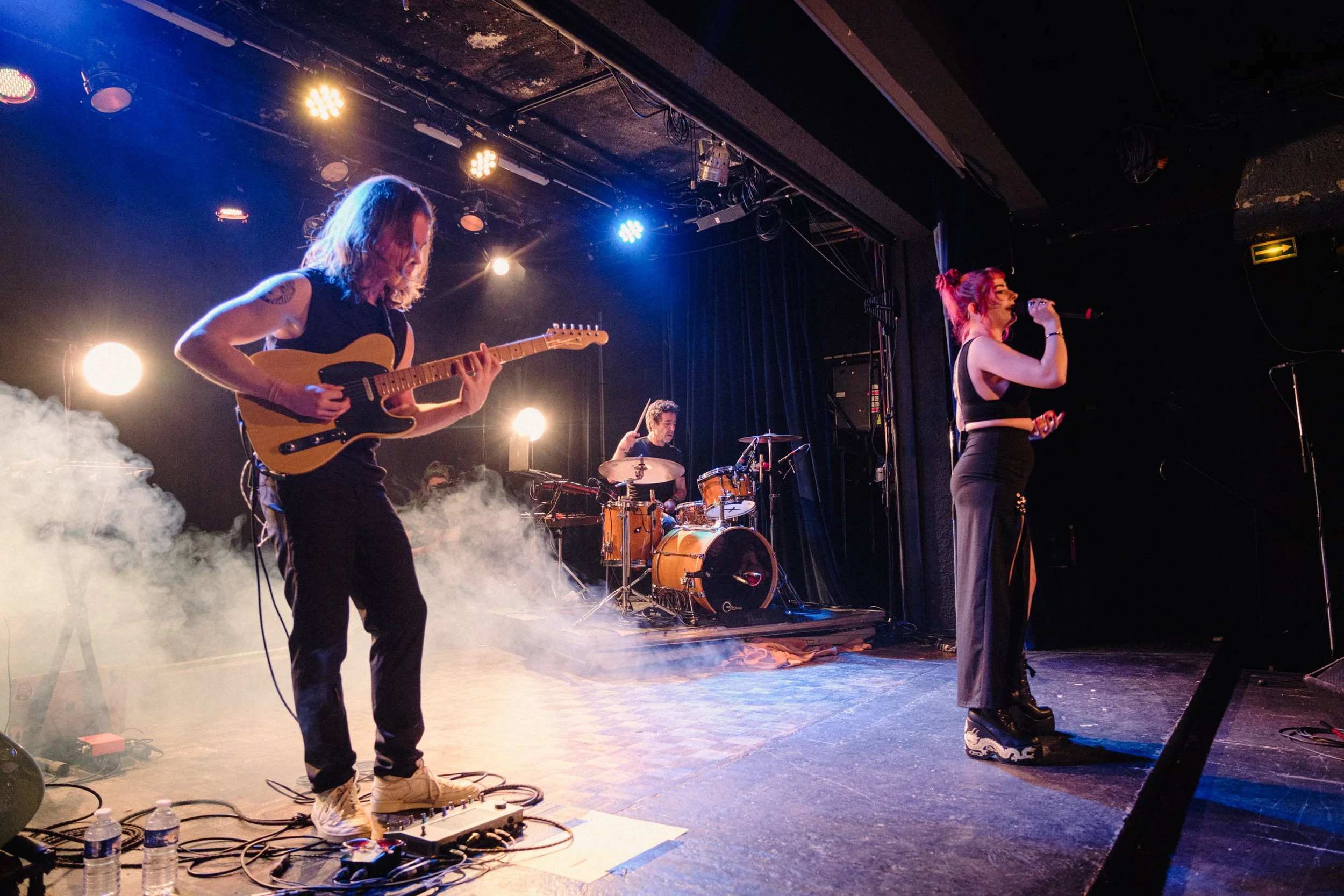 A band performing on stage, with a guitarist on the left, a drummer in the center, and a female singer on the right, with stage lighting and smoke effects.