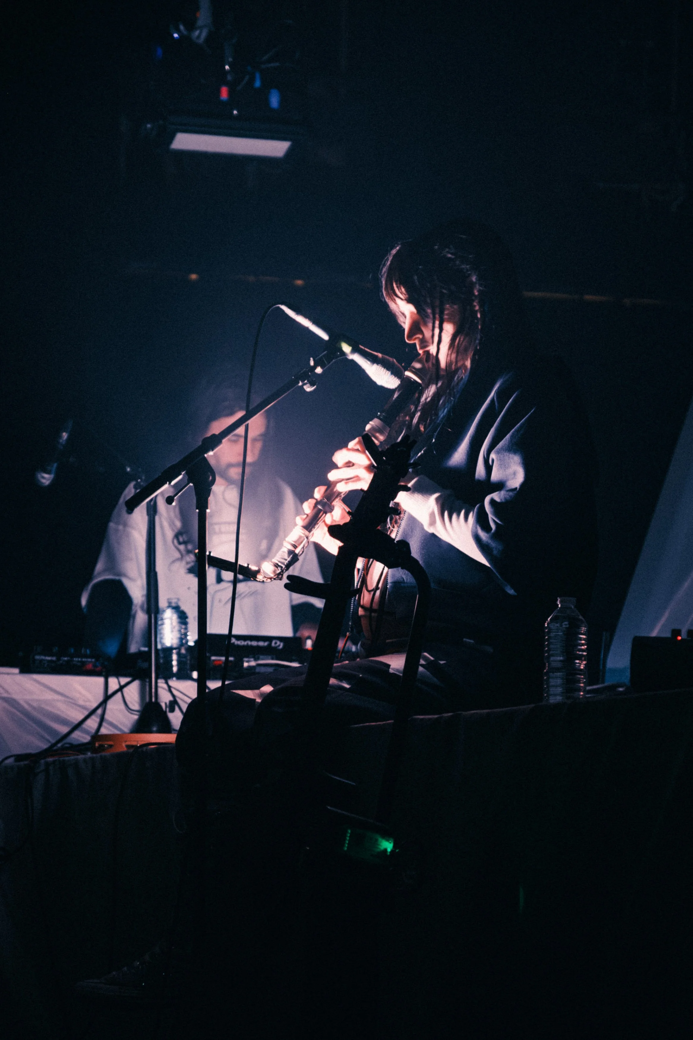 A musician playing a wooden flute on stage during a live performance, with a DJ setup and water bottles nearby, under dark lighting.