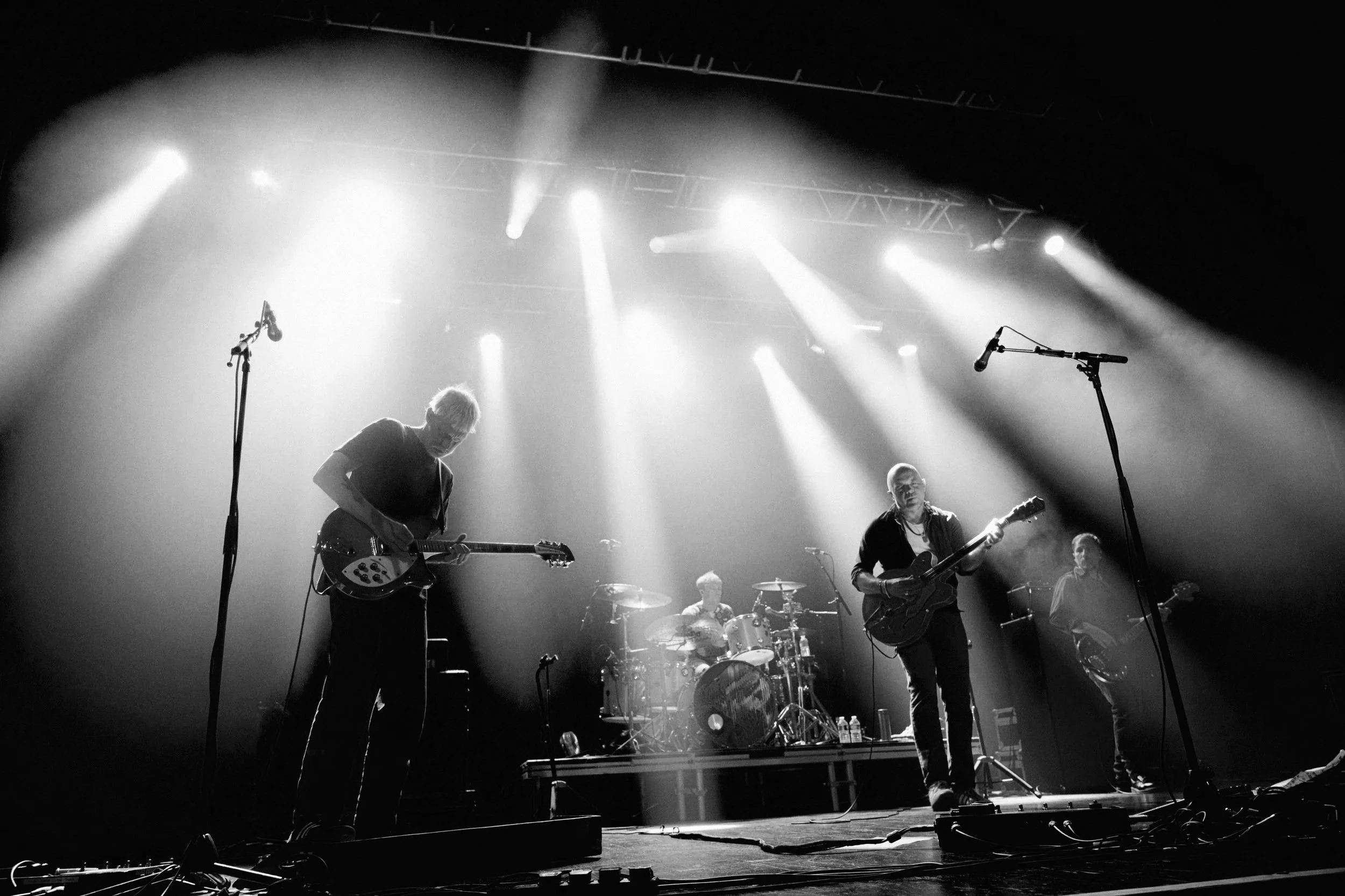 Black and white photo of a four-member band performing on stage with bright lights overhead, playing guitars and drums, with microphones and cables in front.