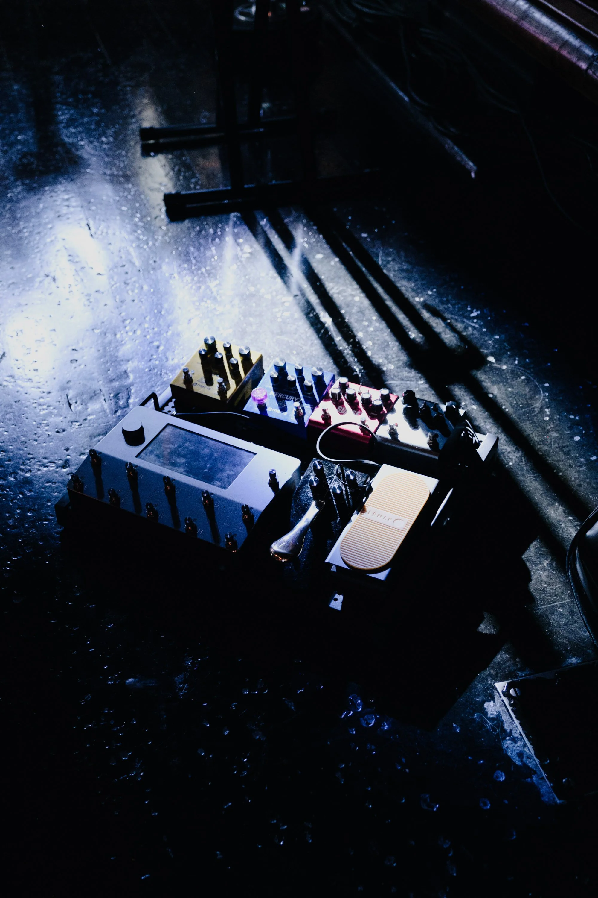 A music pedalboard with various effects pedals and a looper pedal on a dark, wet floor with reflections and shadows.