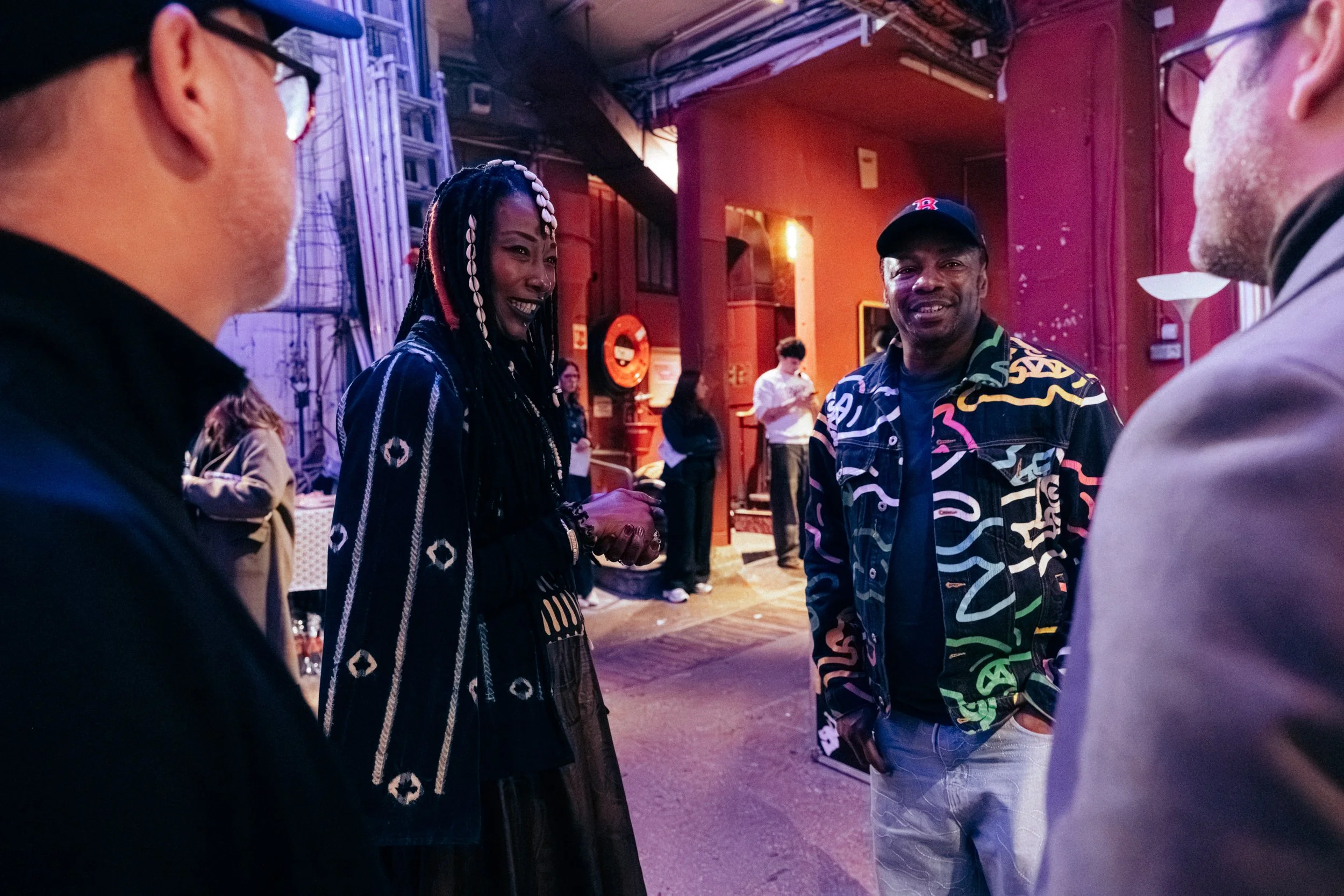 Four people are having a conversation in a lively indoor setting with colorful lighting. One woman is smiling and holding hands with a man, while another man and an additional person are partially visible in the foreground.