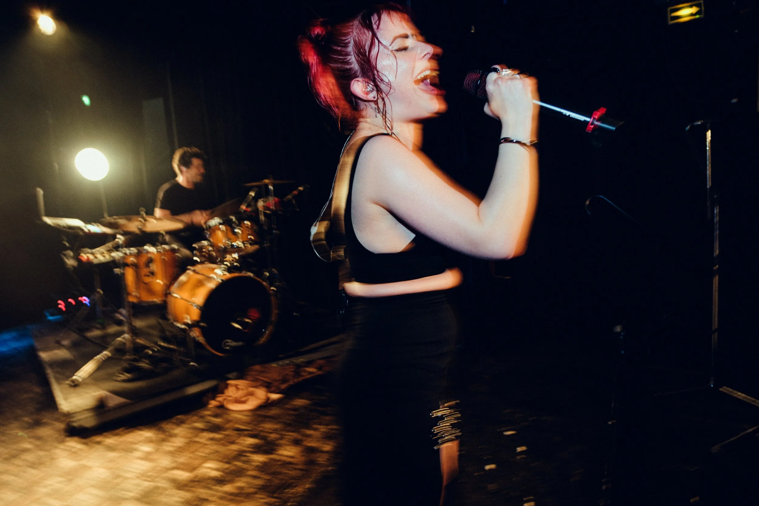 A female singer with red hair singing into a microphone on stage with a drummer playing behind her; dark stage with lighting.