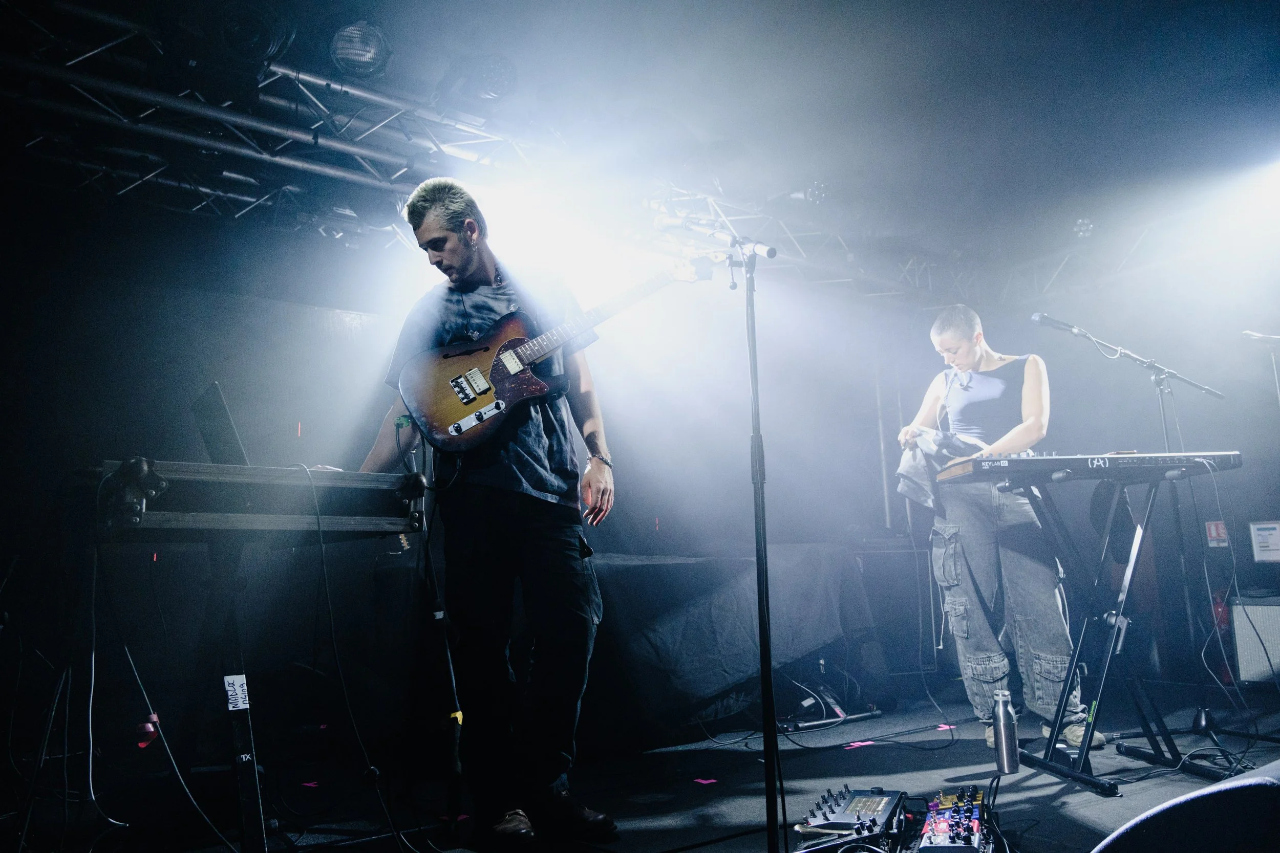 Musicians performing on stage with guitars and keyboard, illuminated by bright stage lighting, with audio equipment and cables visible.