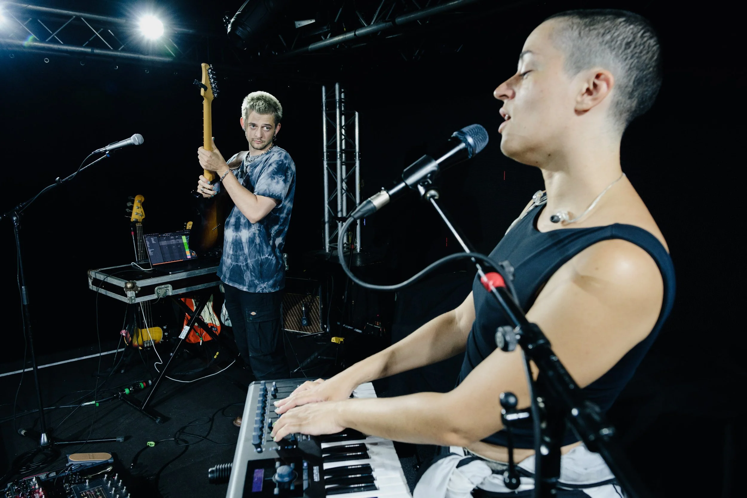 Two musicians performing on stage, one playing keyboard and the other holding an electric guitar, with various electronic equipment around them.