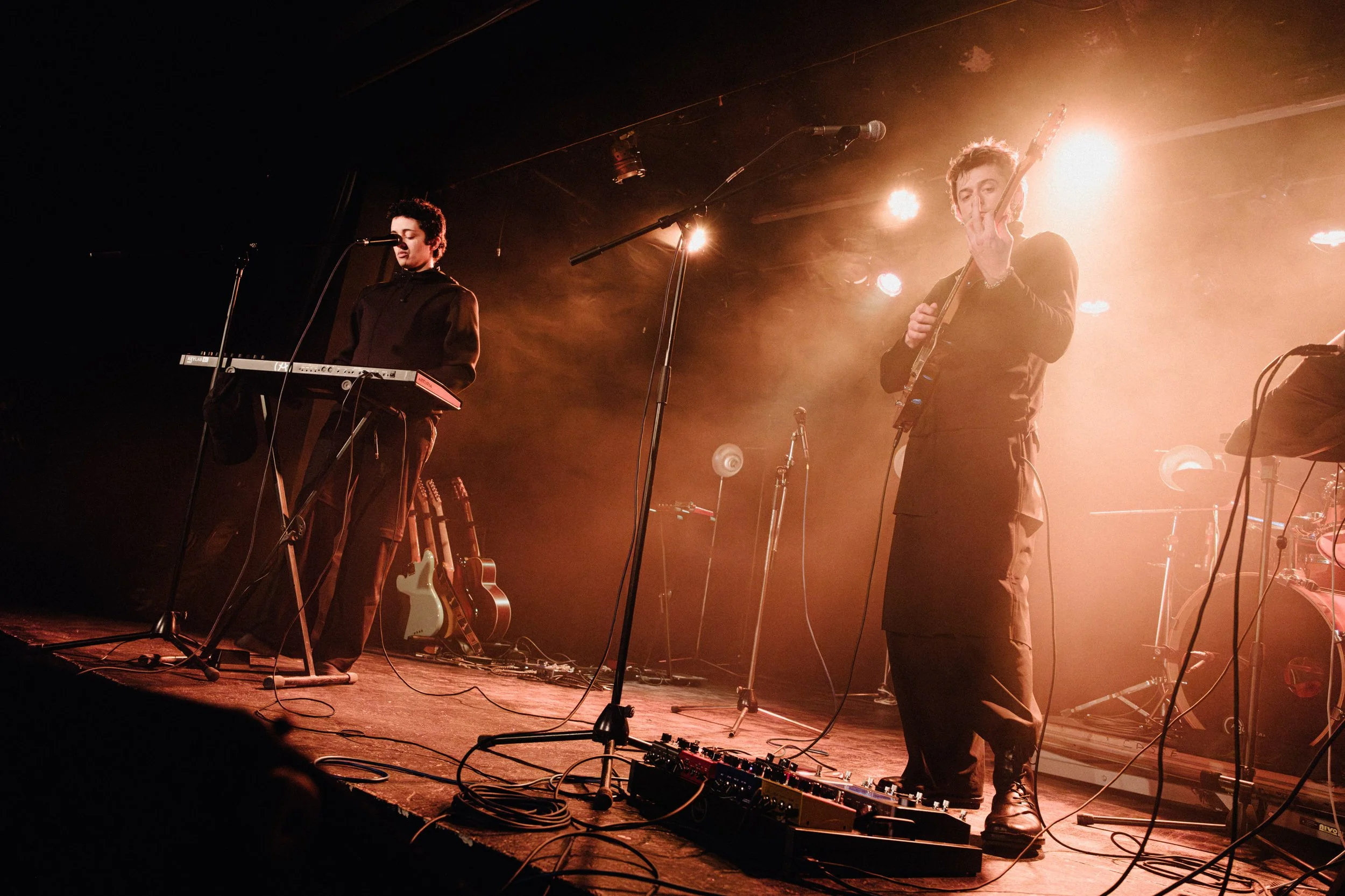 Two musicians on stage performing under warm lighting, one playing keyboard and singing into a microphone, the other playing guitar and singing, with guitar amplifiers and multiple guitars visible in the background.