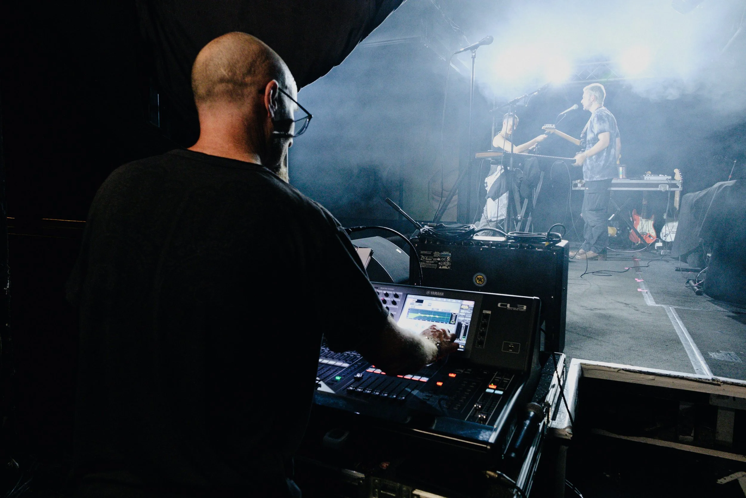 Behind-the-scenes view of a sound engineer working on a mixing console during a live music performance. In the background, musicians perform on stage with bright lighting and musical equipment.