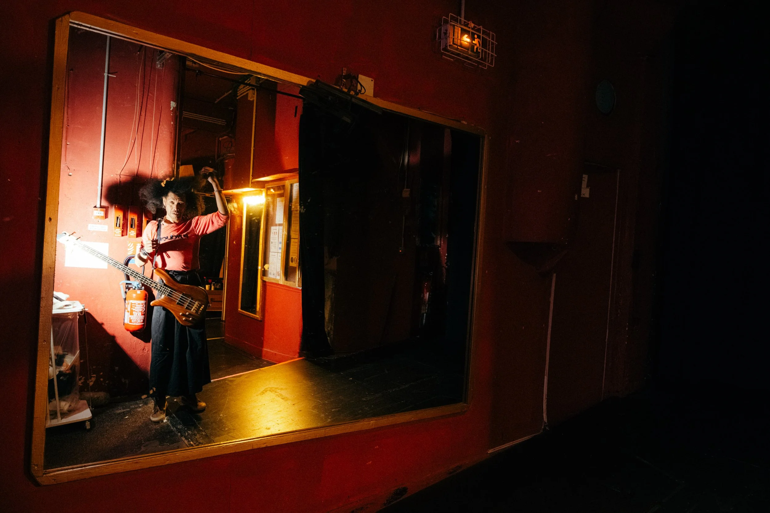 Person standing in backstage area holding a bass guitar, illuminated by warm lighting, with red walls and various equipment around.
