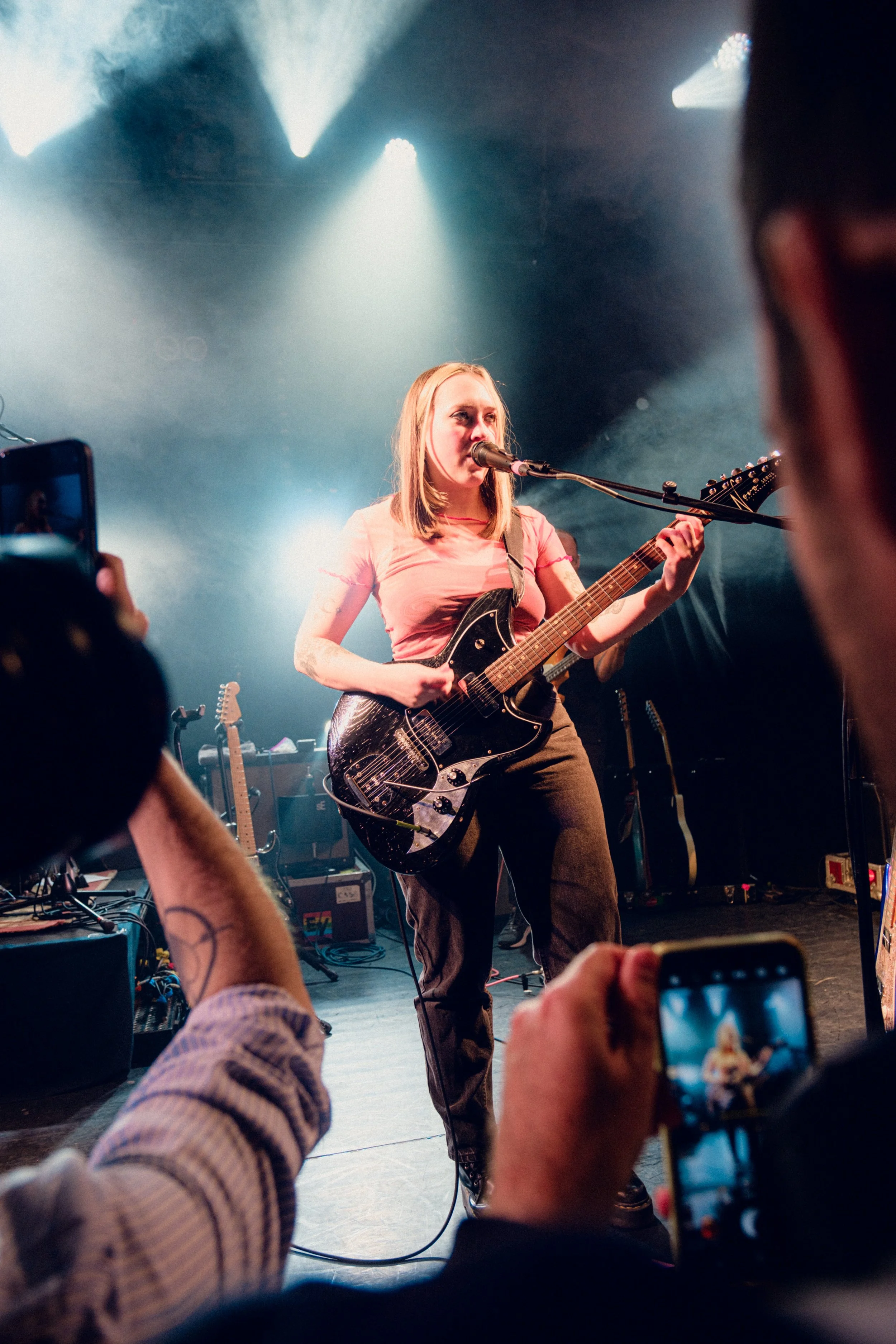 A female musician performing on stage with a guitar, singing into a microphone, illuminated by stage lights, while audience members capture the moment on their phones.