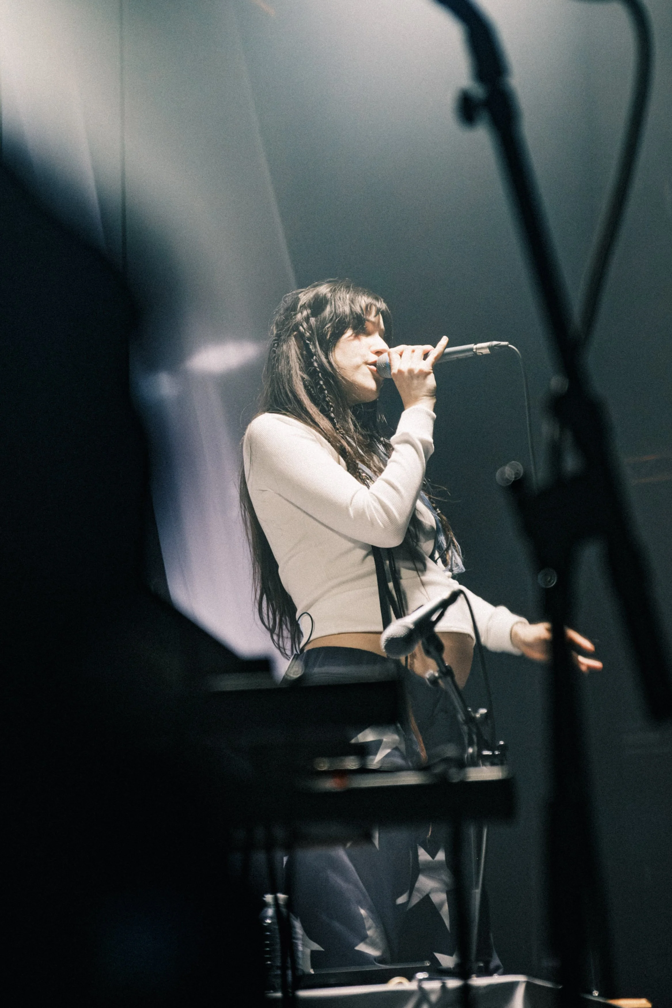 A young woman singing into a microphone on stage, with stage equipment and a dark background.