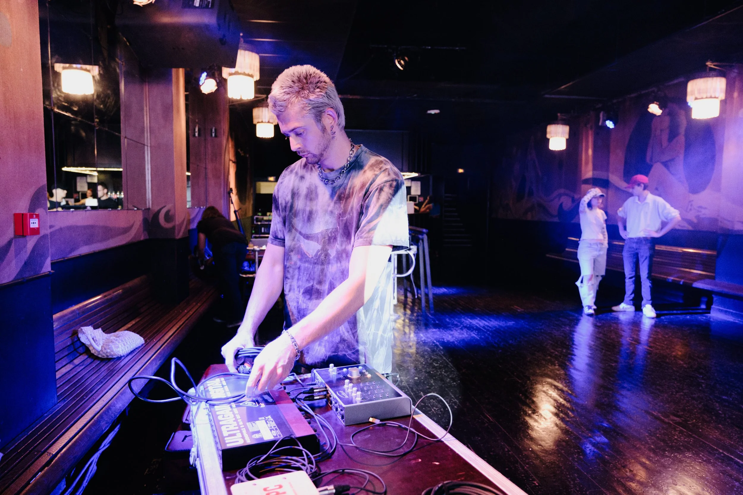 A DJ with platinum blonde hair and tattoos is operating DJ equipment in a dimly lit nightclub. Two people are talking in the background, and the dance floor is empty with a purple-blue lighting ambiance.