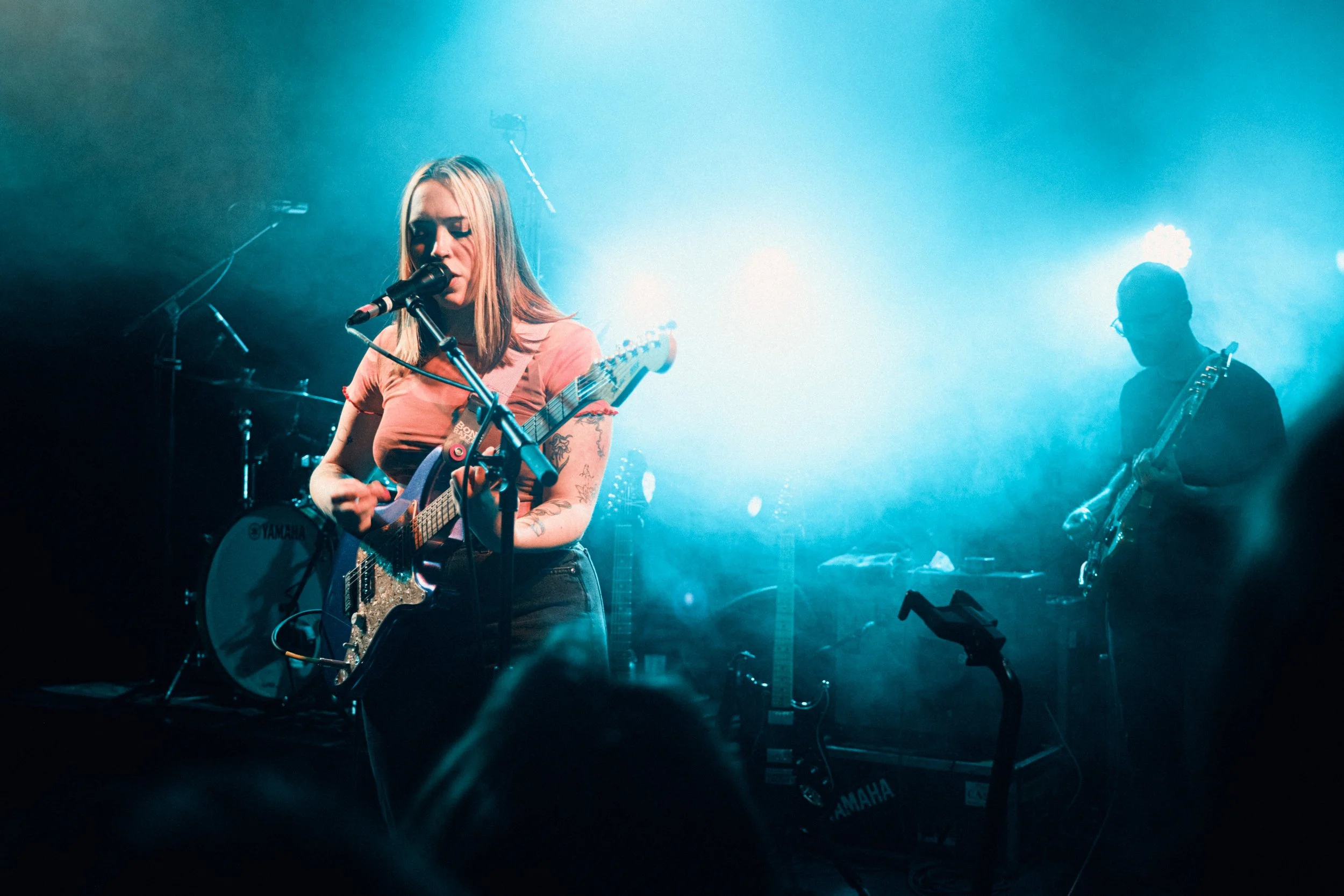 Female musician singing and playing guitar on stage with a male musician playing guitar in the background, stage lights create a blue ambiance.