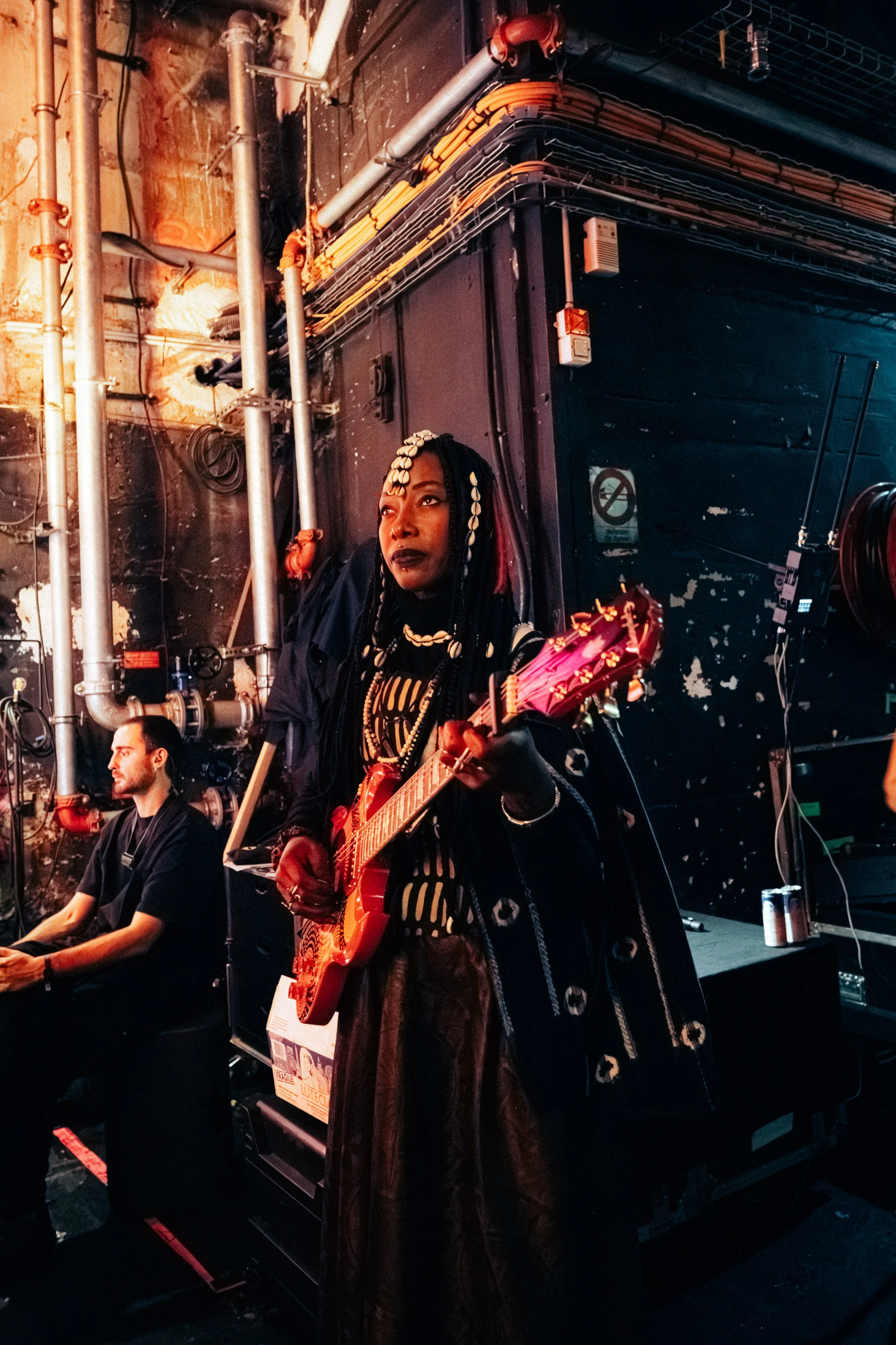 A woman with braided hair adorned with shells, wearing dark clothing, playing a pink electric guitar on stage. A man is sitting beside her with electronic equipment, and the background features a black wall with exposed pipes and electrical wiring.