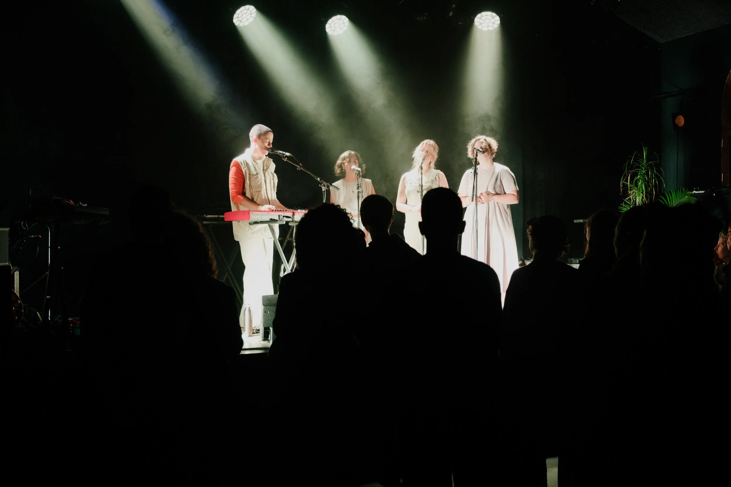 A musical performance on stage with four female singers and one keyboard player, lit by spotlights, audience members silhouetted in the foreground.