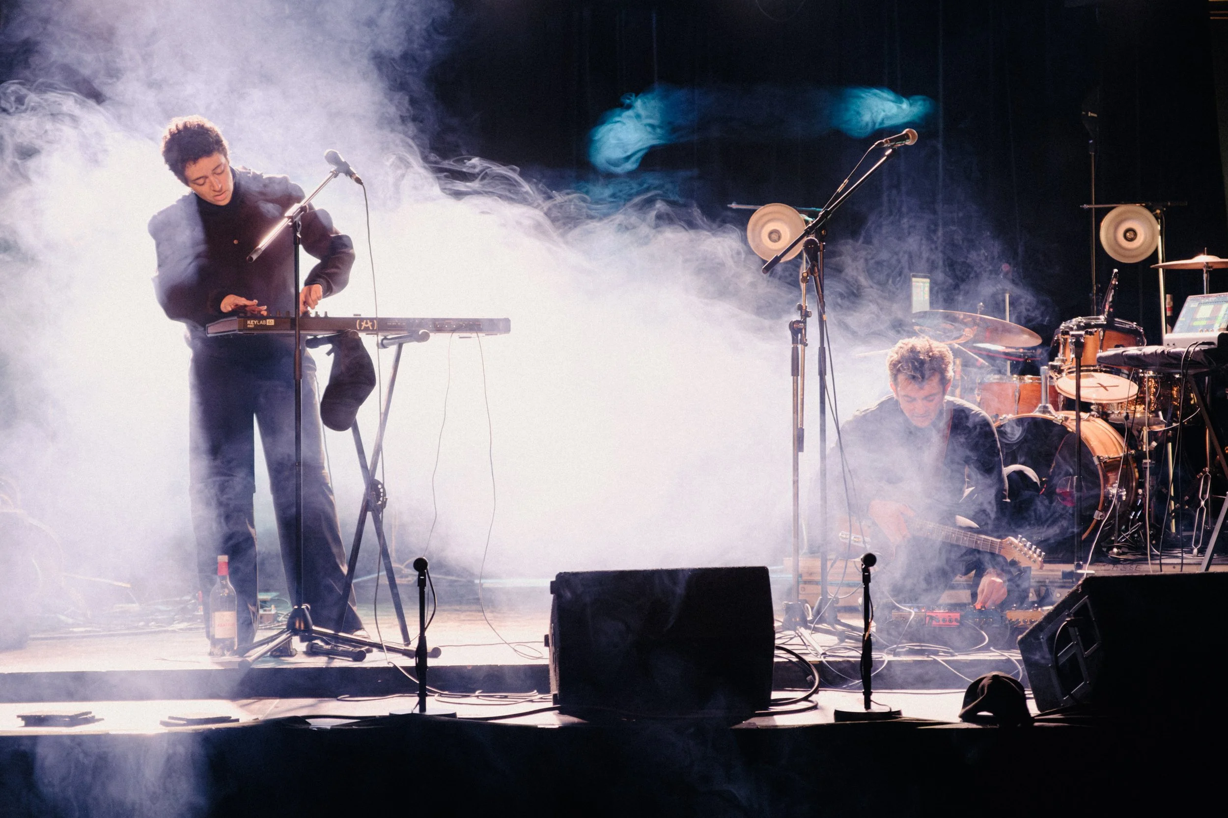 Two musicians performing on stage with fog effects. One standing and playing a keyboard, the other sitting and playing an electric guitar, with drum set and microphones visible.
