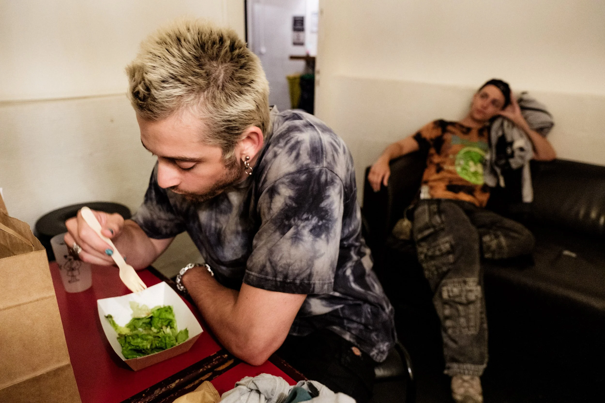 A young man with blonde hair eating a salad from a takeout container while sitting at a table. In the background, a woman with dark hair is lounging on a couch, resting her head on a pillow.