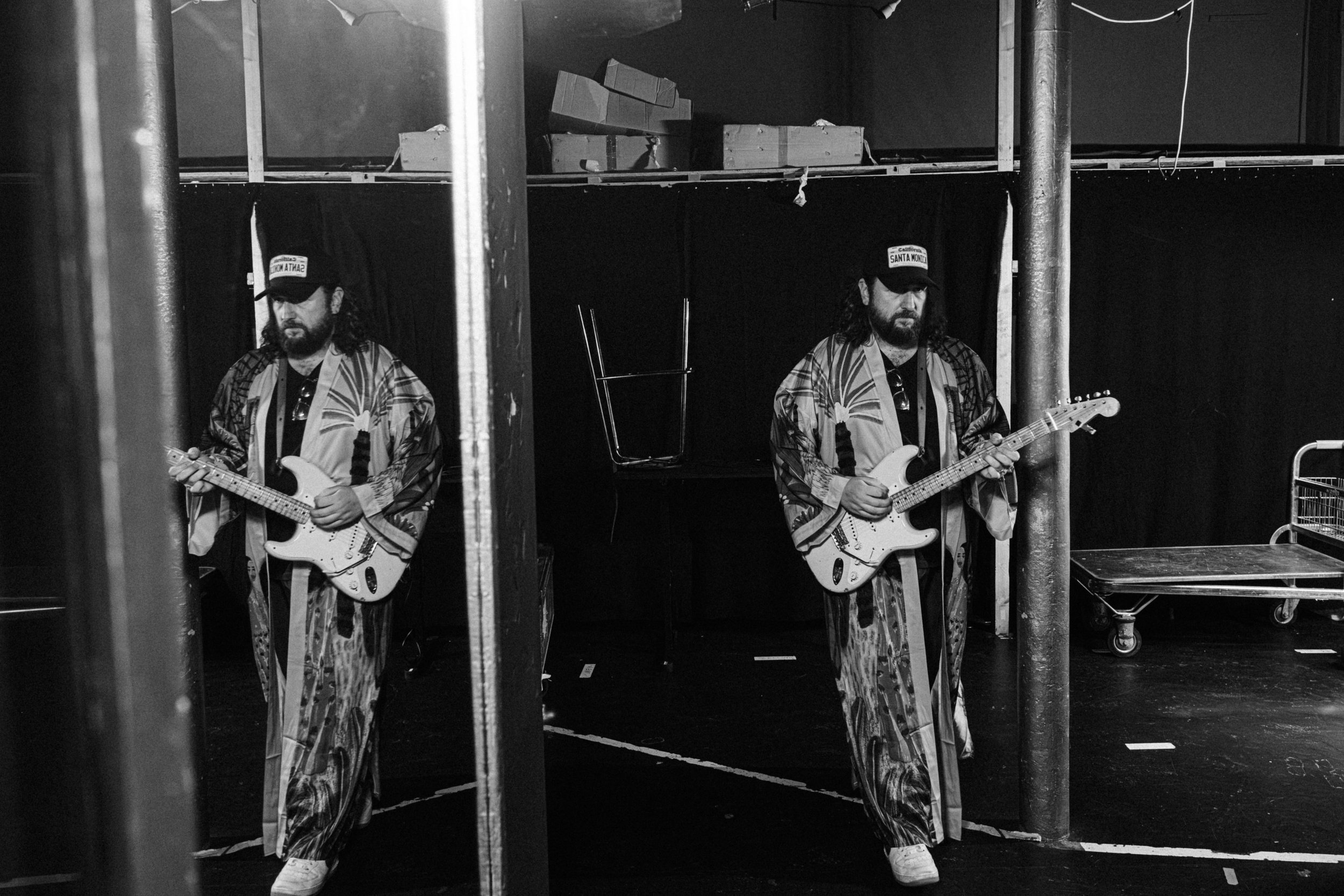 Two men standing in a backstage area with black curtains, each holding an electric guitar. They are dressed in patterned clothing and wearing caps that say "Santa Monica." The area has some shelving and a rolling cart, with one person reflected in th