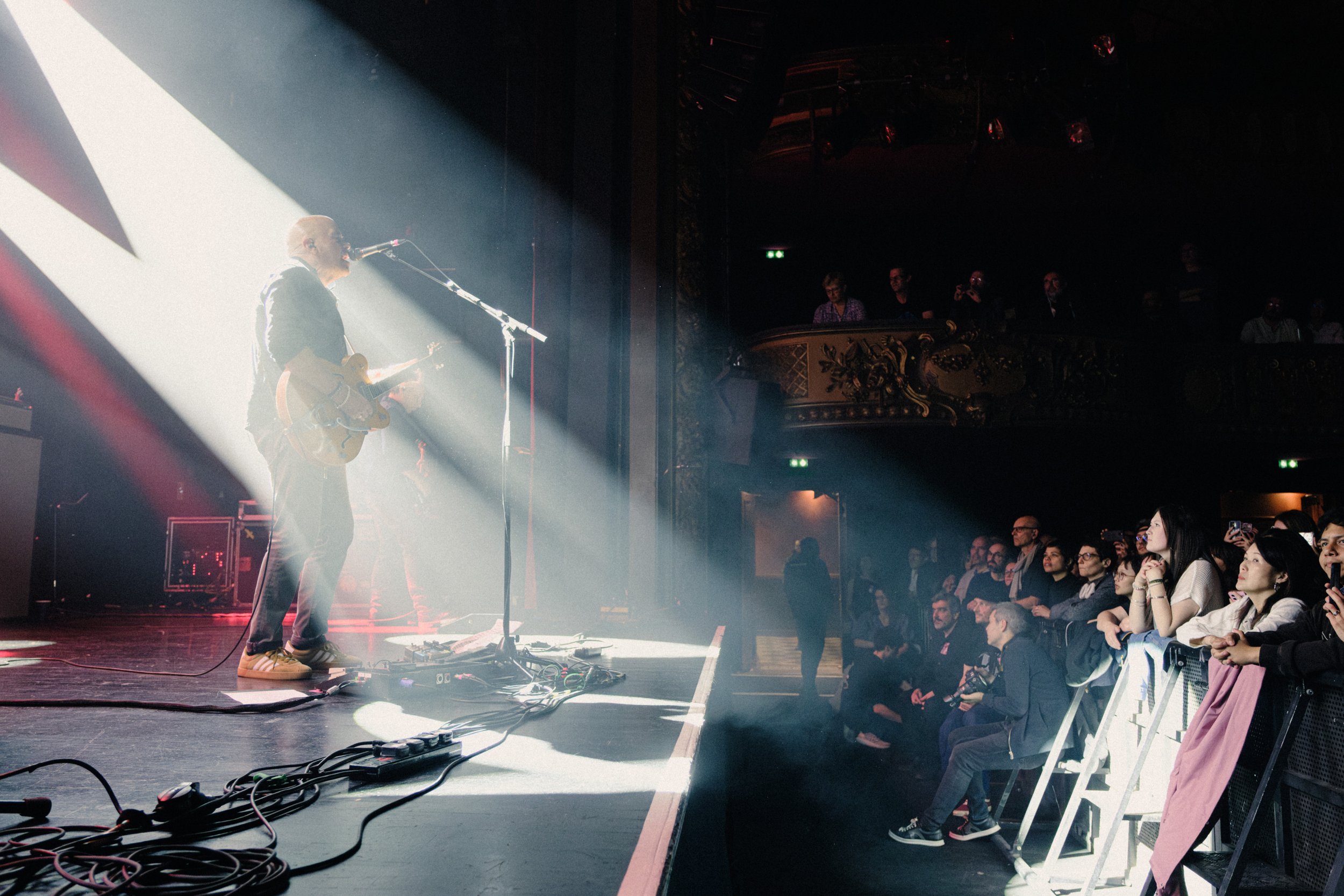 A band performing on stage with one musician playing a guitar and singing into a microphone, illuminated by a bright spotlight. Audience members are seated and standing in front of the stage, watching the performance in a dark concert venue.