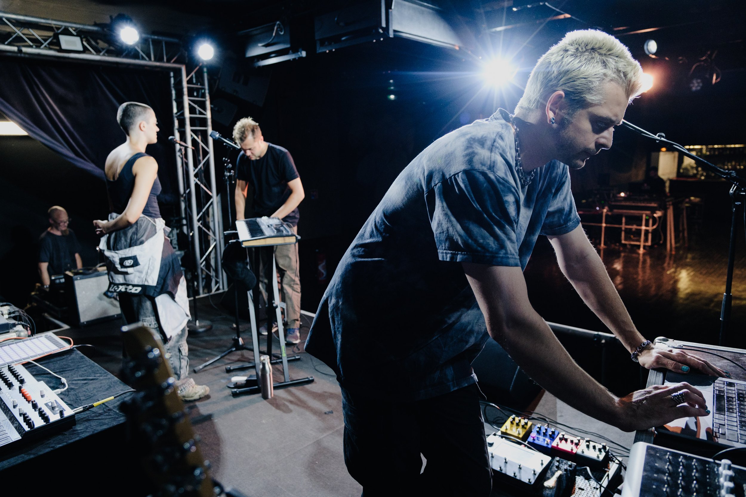 Musicians performing on stage with electronic equipment and instruments during a live show, with a bright stage light overhead.