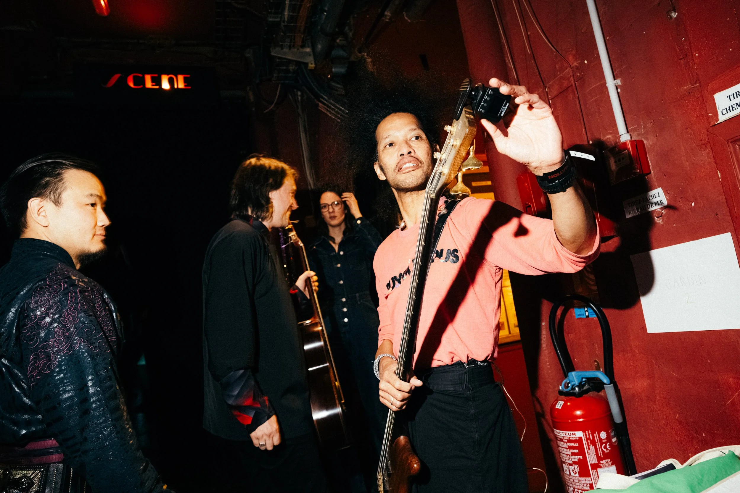 A group of people, including a man with an afro holding a guitar, are backstage preparing for a performance in a dimly lit venue with red walls. They are engaging with each other, and some are holding instruments.