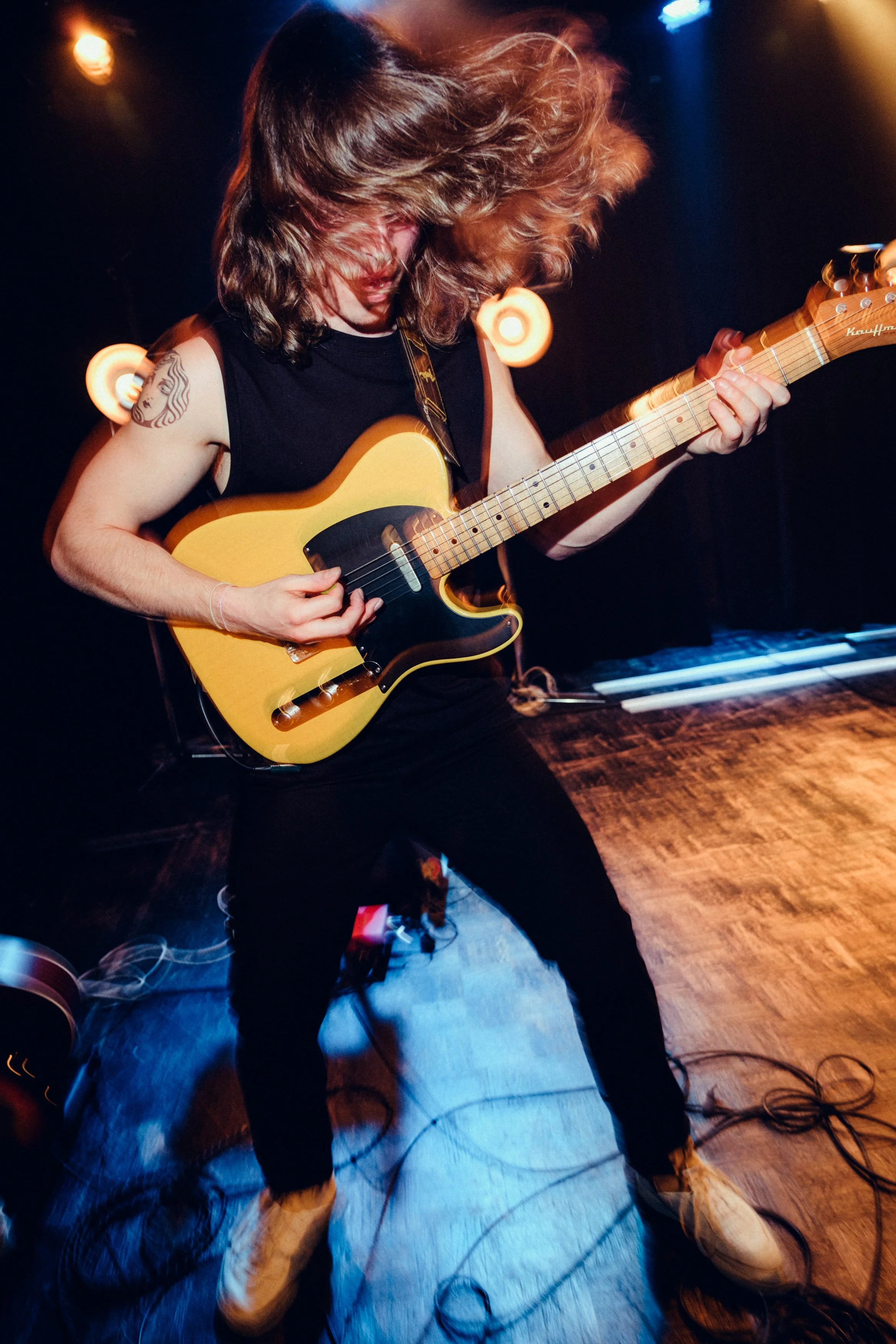 A person with long hair playing an electric guitar on stage, with motion blur from their hair and movement.