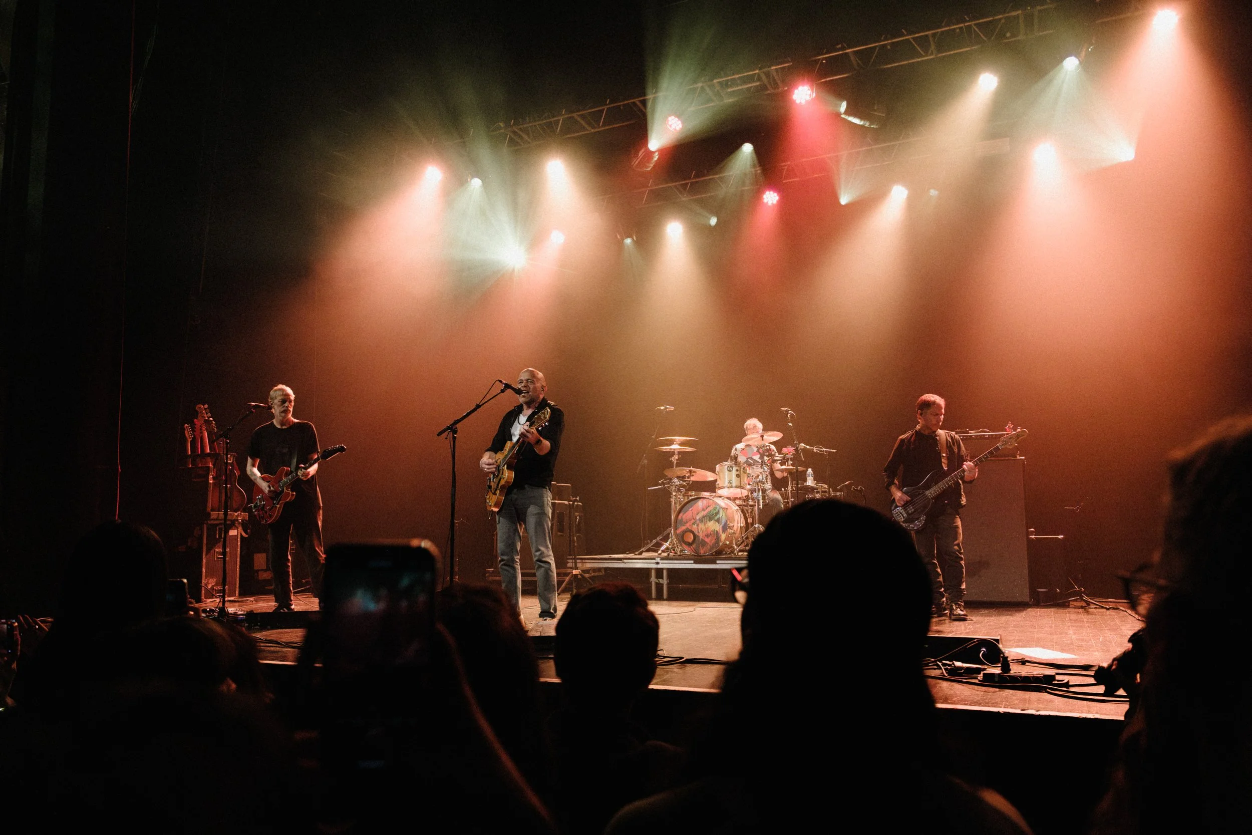 A band performing live on stage with four members playing guitars, drums, and bass, under colorful stage lighting, with audience members in the foreground