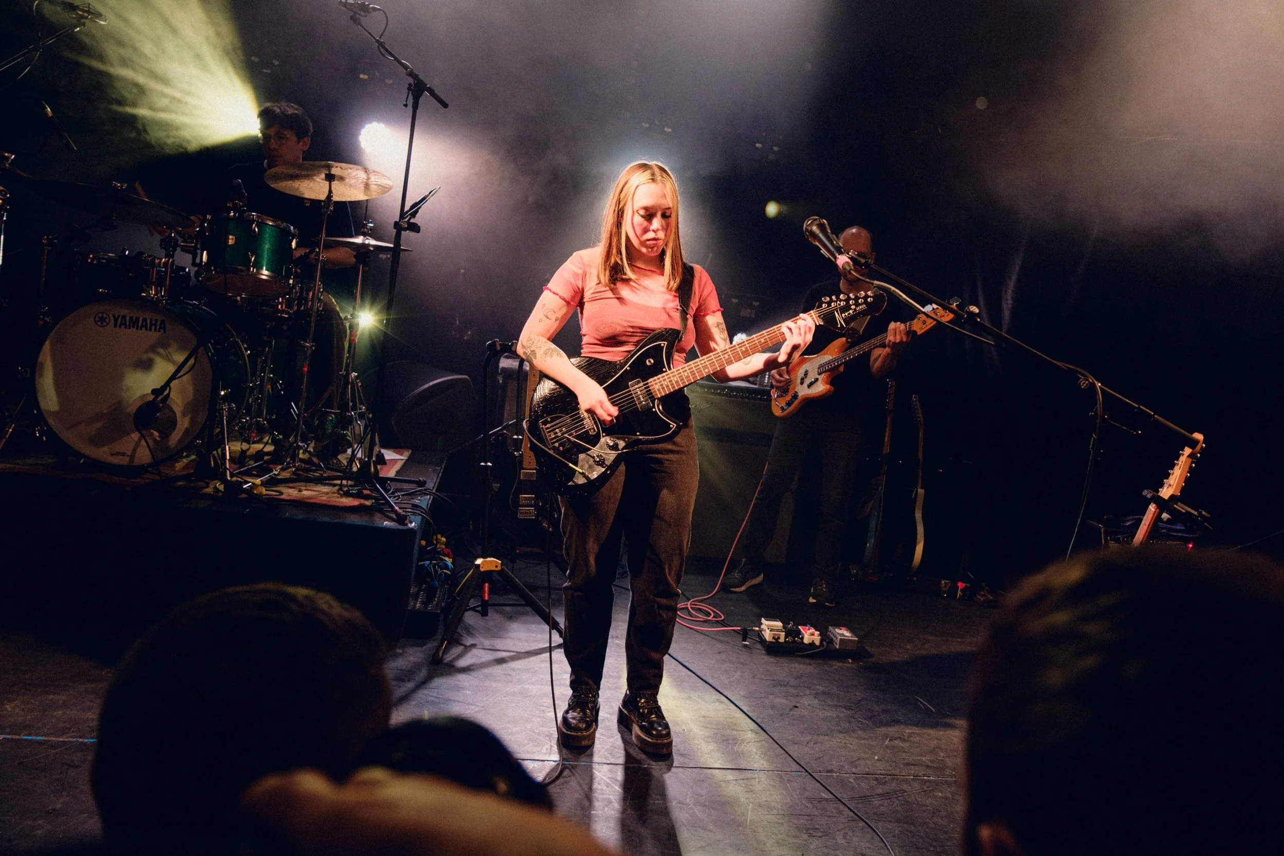 A female musician plays an electric guitar on stage during a concert, with a drummer and another guitarist in the background, and stage lights illuminating the scene.