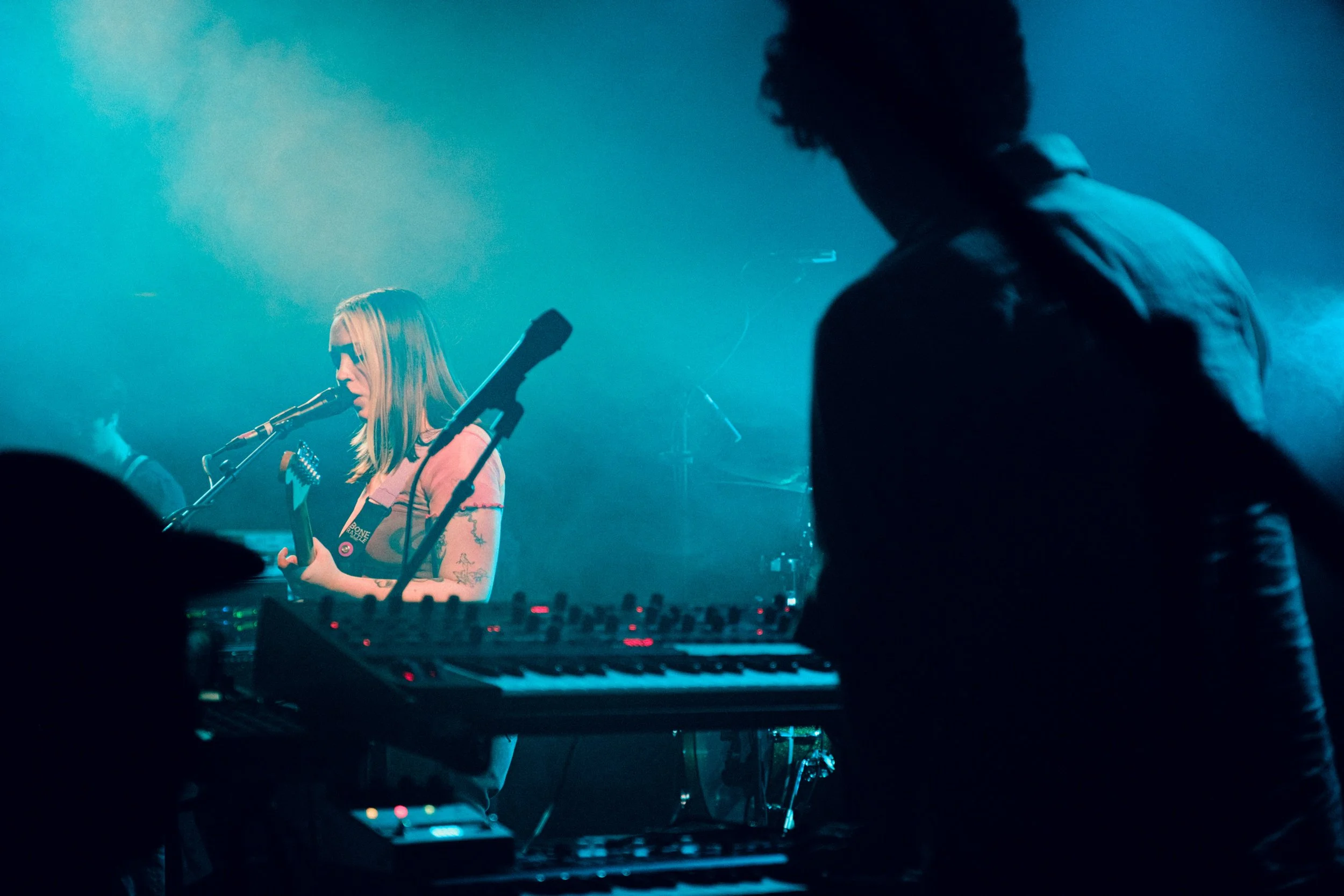 Female musician singing into a microphone while playing guitar on stage with blue lighting, and male musician in the foreground playing keyboard.