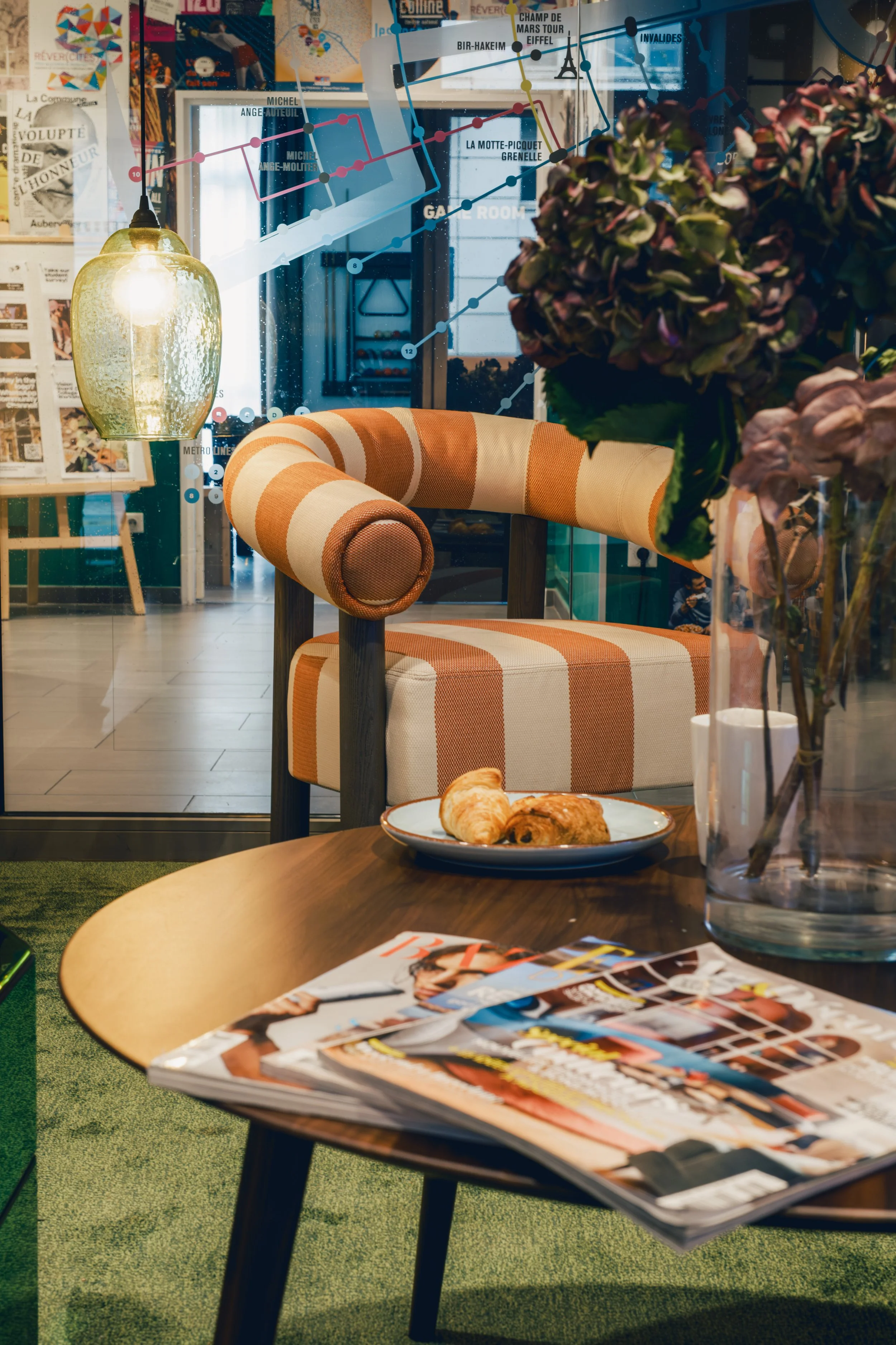 A cozy interior space with a striped armchair, a wooden coffee table with magazines and pastries, a large vase with flowers, and a glass partition with a cityscape and graph overlay on the window.