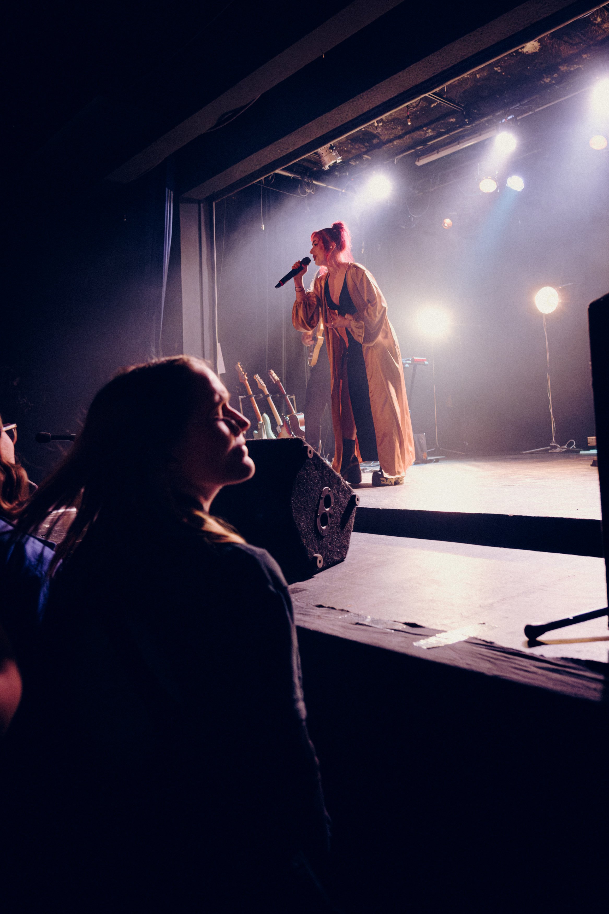 A woman with pink hair singing into a microphone on stage during a performance, illuminated by stage lights, with audience members watching.