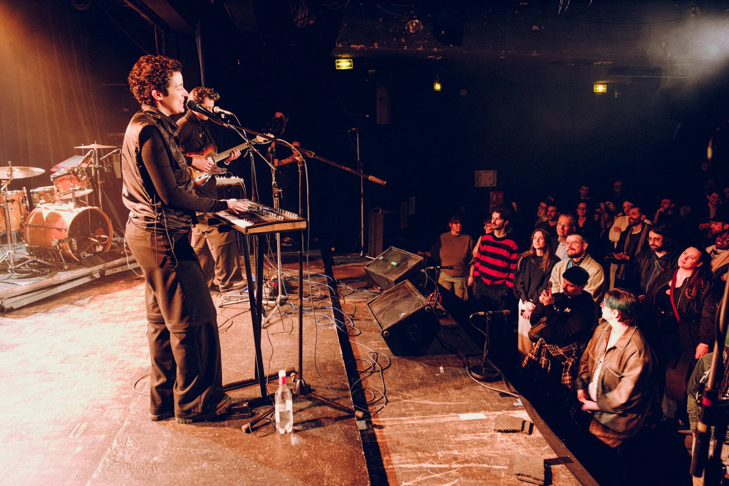 Musicians perform on stage with instruments including drums and keyboard, facing a standing crowd in a dimly lit concert venue.