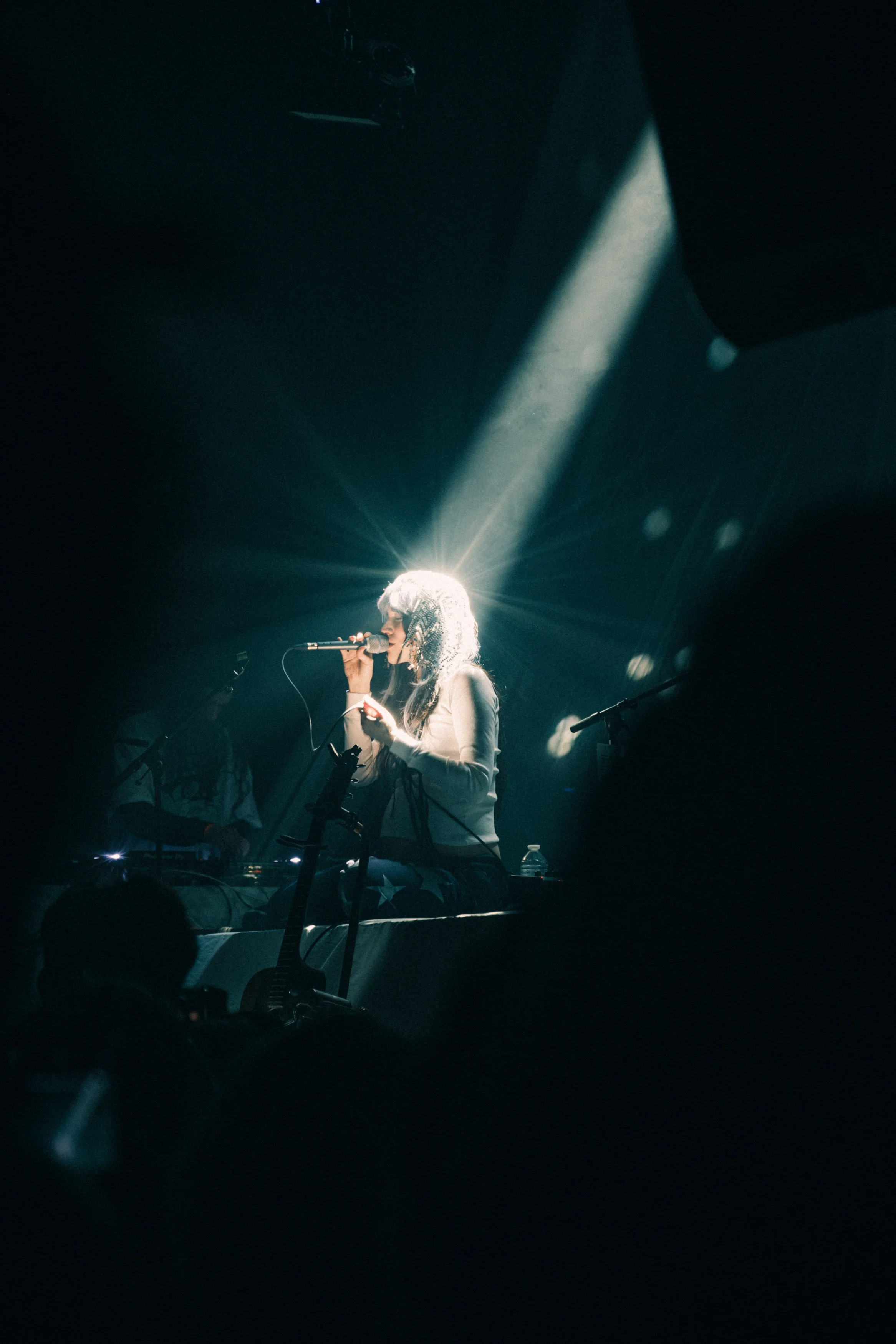 A female singer performing on stage with a microphone, illuminated by a bright light behind her, creating a starburst effect, with a DJ or technician visible in the background.