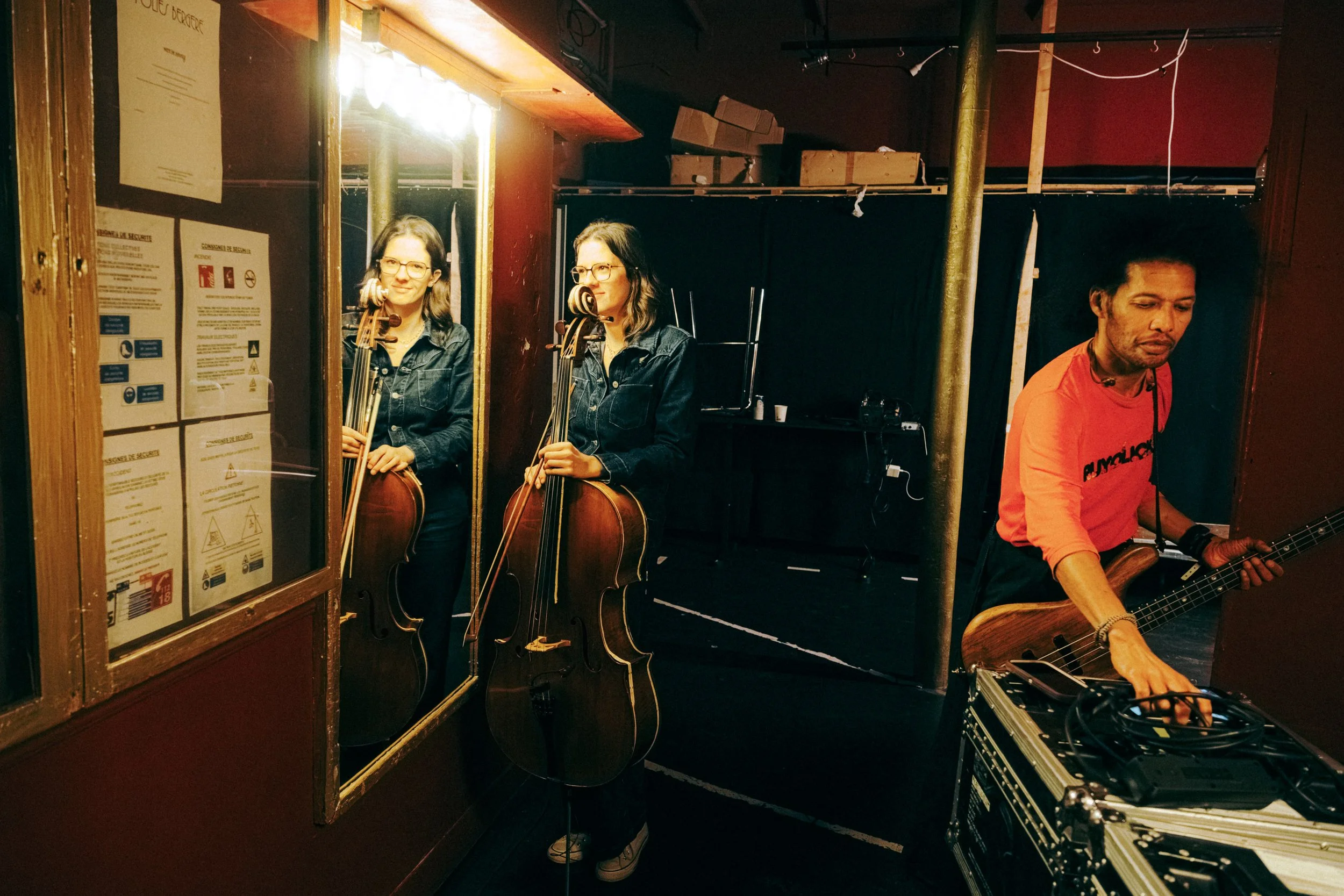 Two women playing double basses are seen through a window reflection, and a man on the right is holding a guitar and some electronic equipment behind a barrier in a dark room, possibly a rehearsal space or stage.
