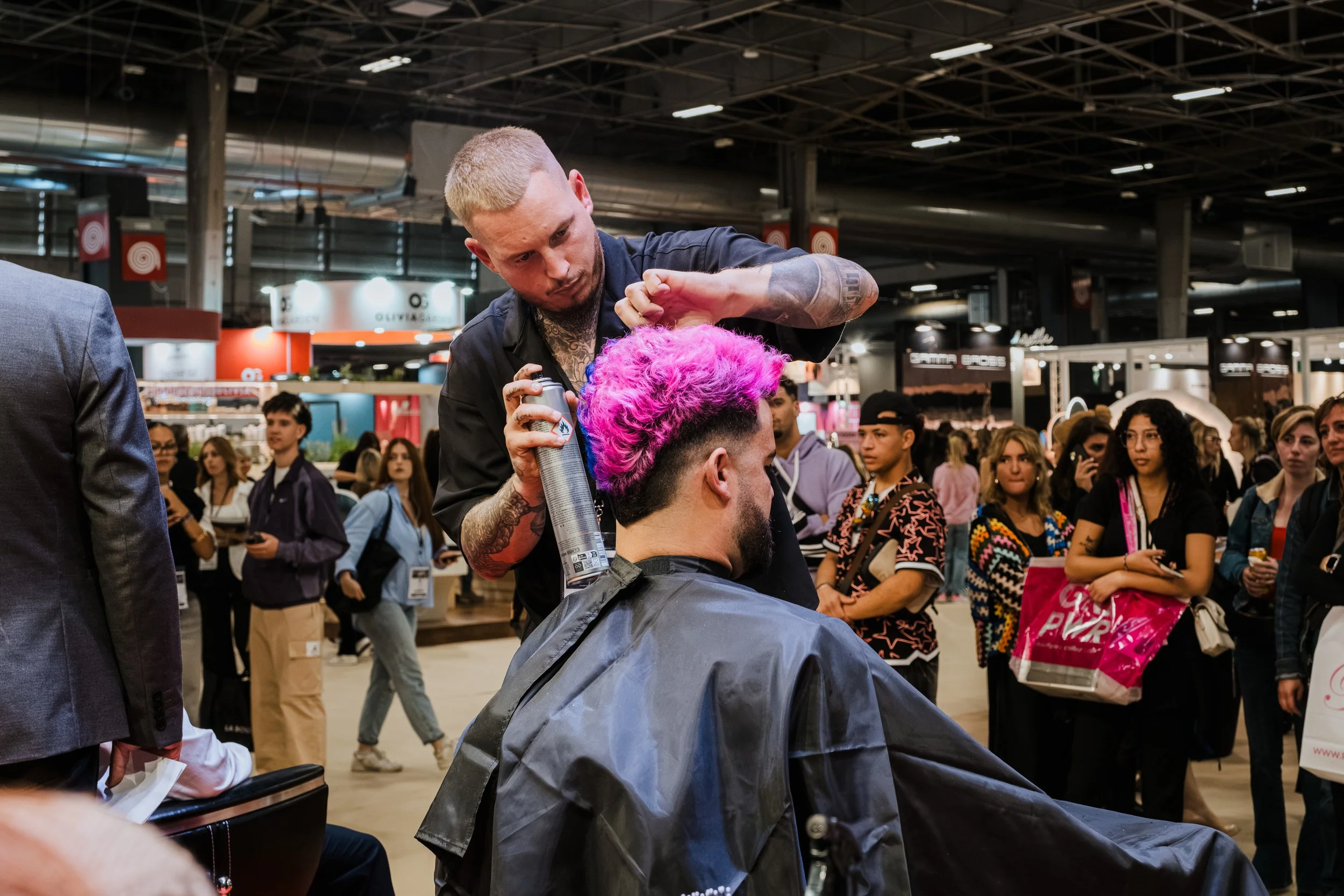 Barber spraying hair of a man with vibrant pink hair in a busy indoor setting, surrounded by onlookers.